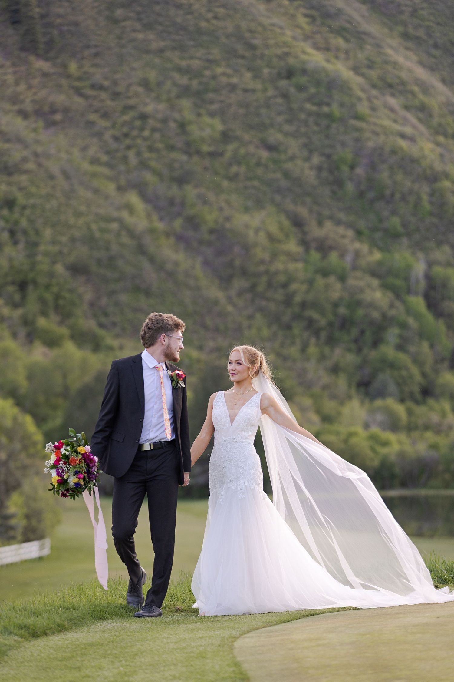 A romantic couple in formal wedding attire walks along a scenic mountain lakeside path at sunset.