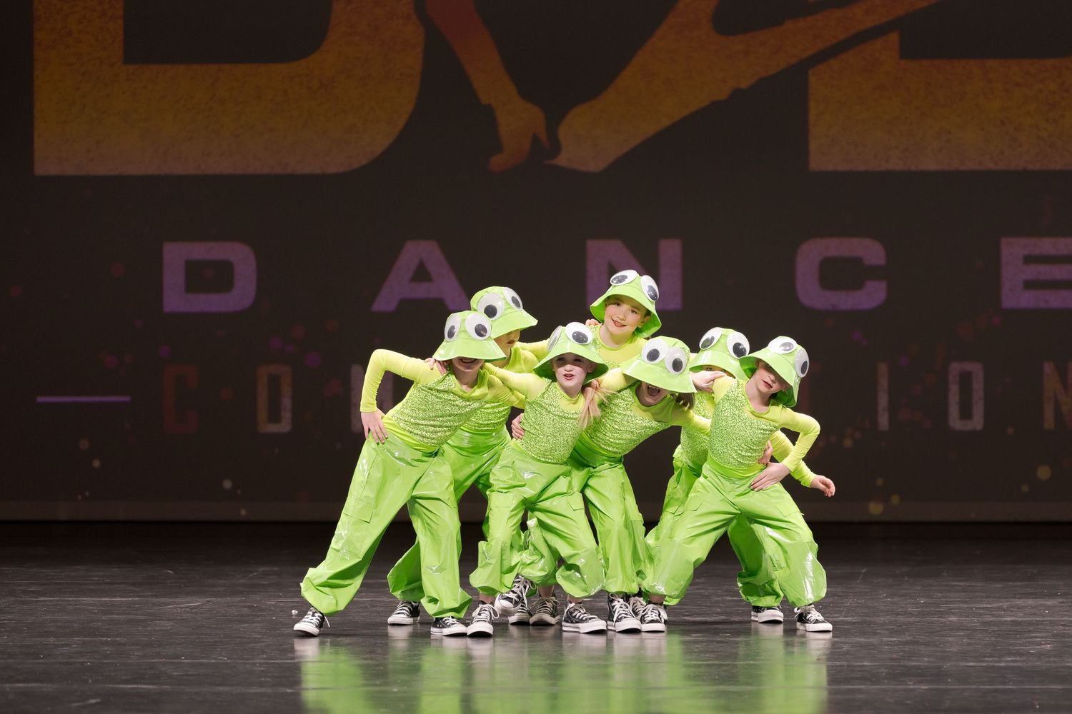 Dancers in bright lime green costumes perform a synchronized hip hop routine on a dark stage.