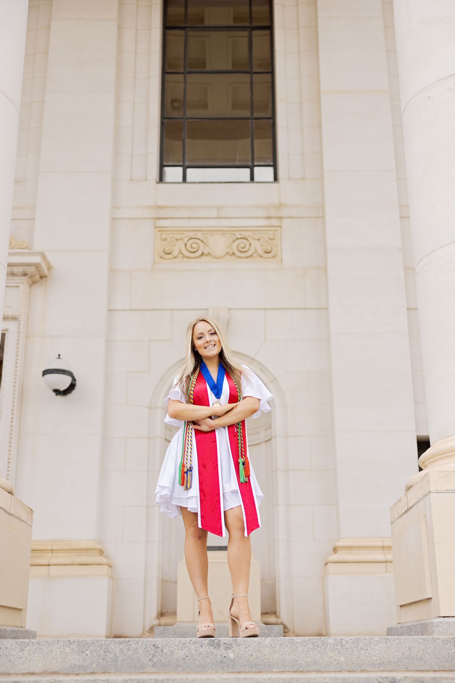 Graduate in white dress and red stole stands confidently on stone steps of building.