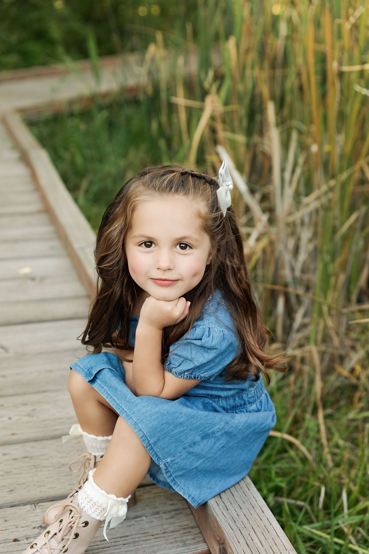 Small child in blue dress sitting on wooden boardwalk surrounded by tall grass and cattails.