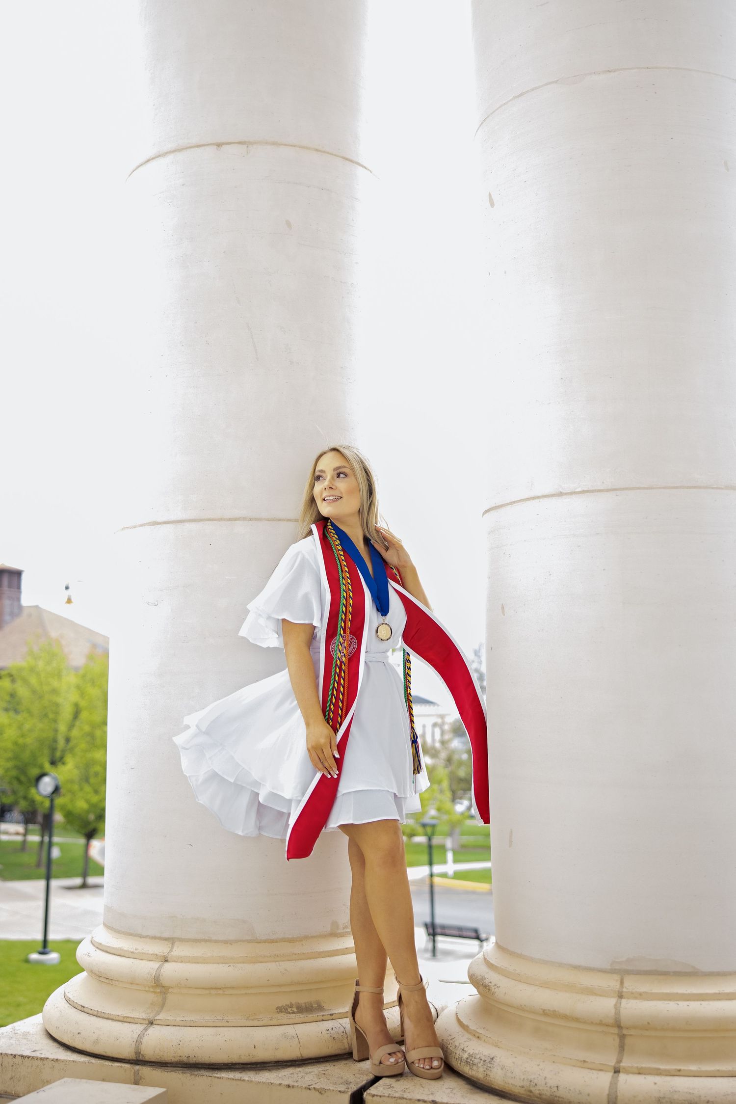 Graduate in white dress and red stole poses between large white columns outdoors.