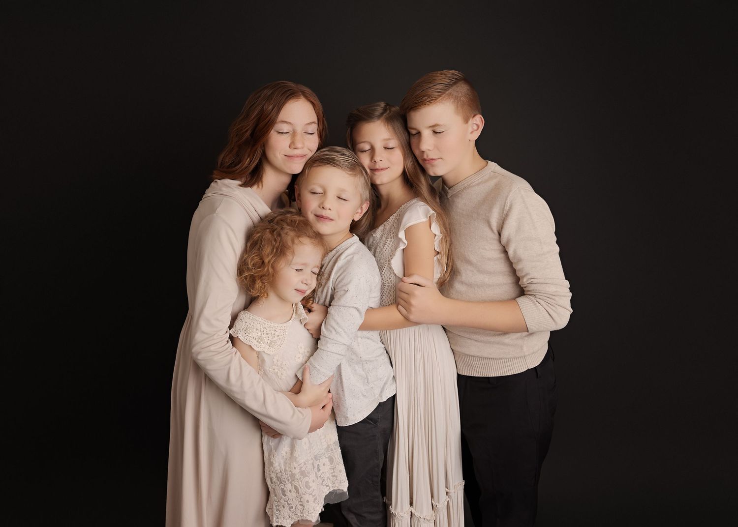 Intimate family portrait with everyone embracing in neutral tones against dark background.