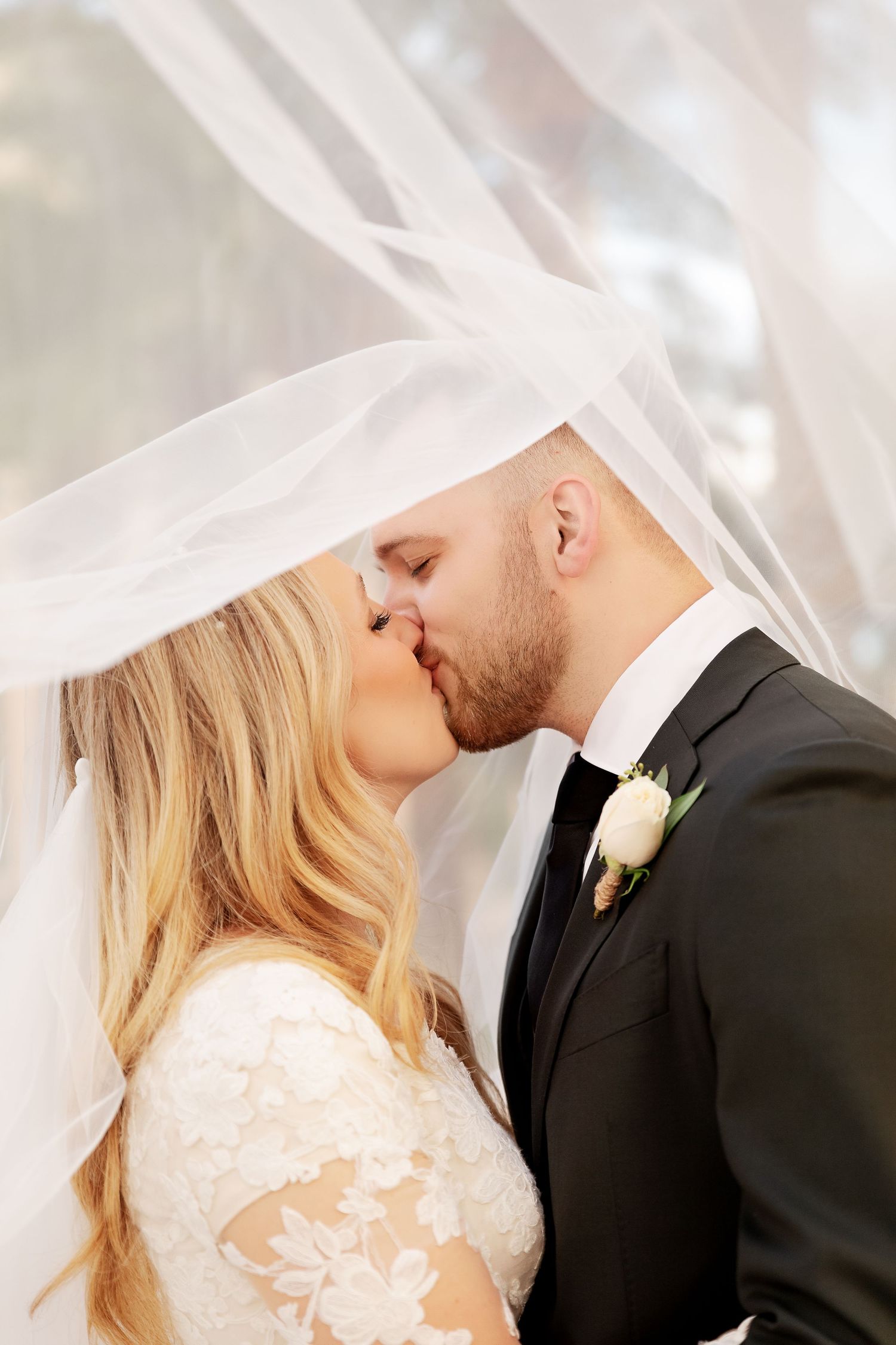 Romantic wedding kiss under flowing veil with white rose boutonniere detail.
