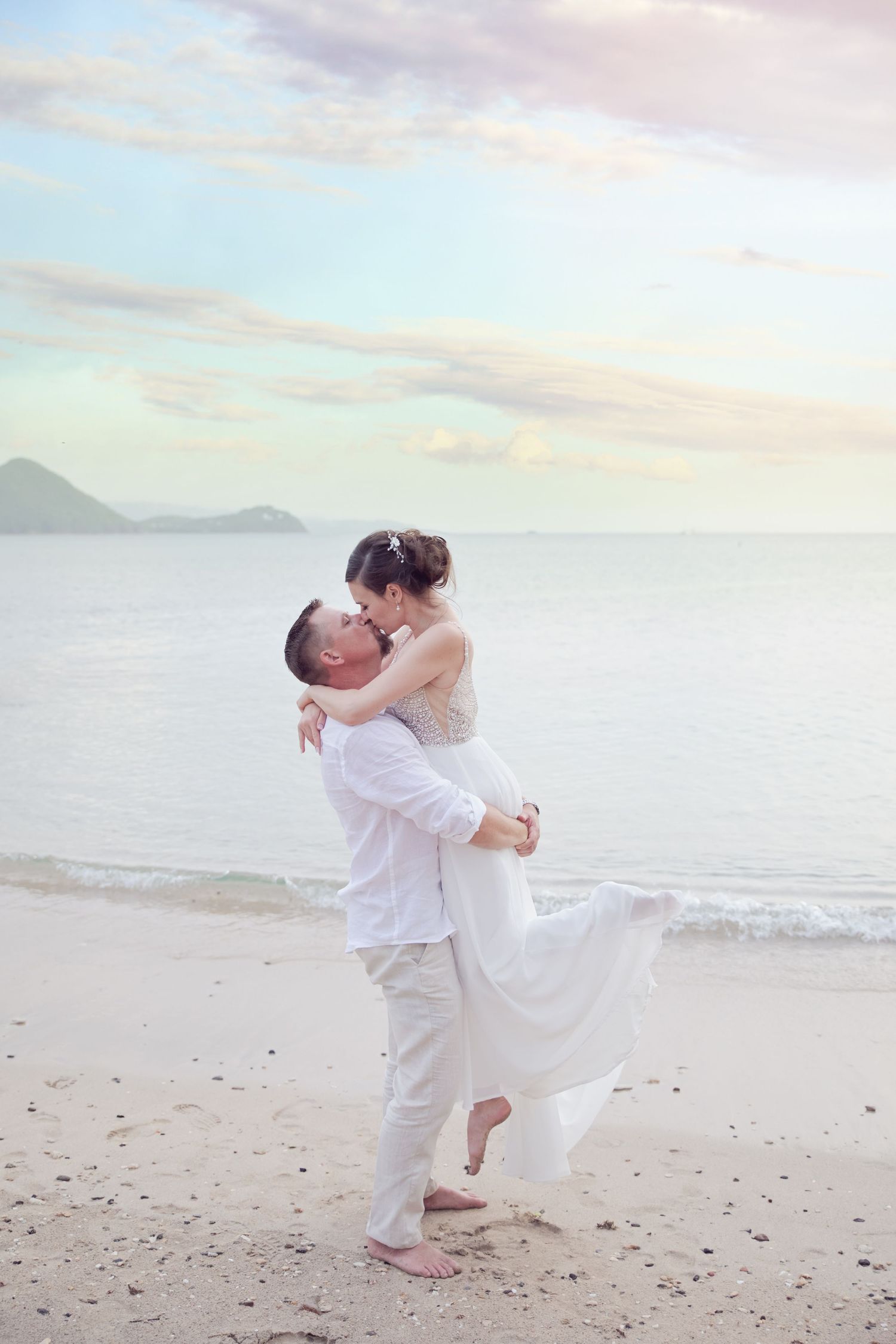 A couple in white attire embracing on a beach with mountains visible in the background.
