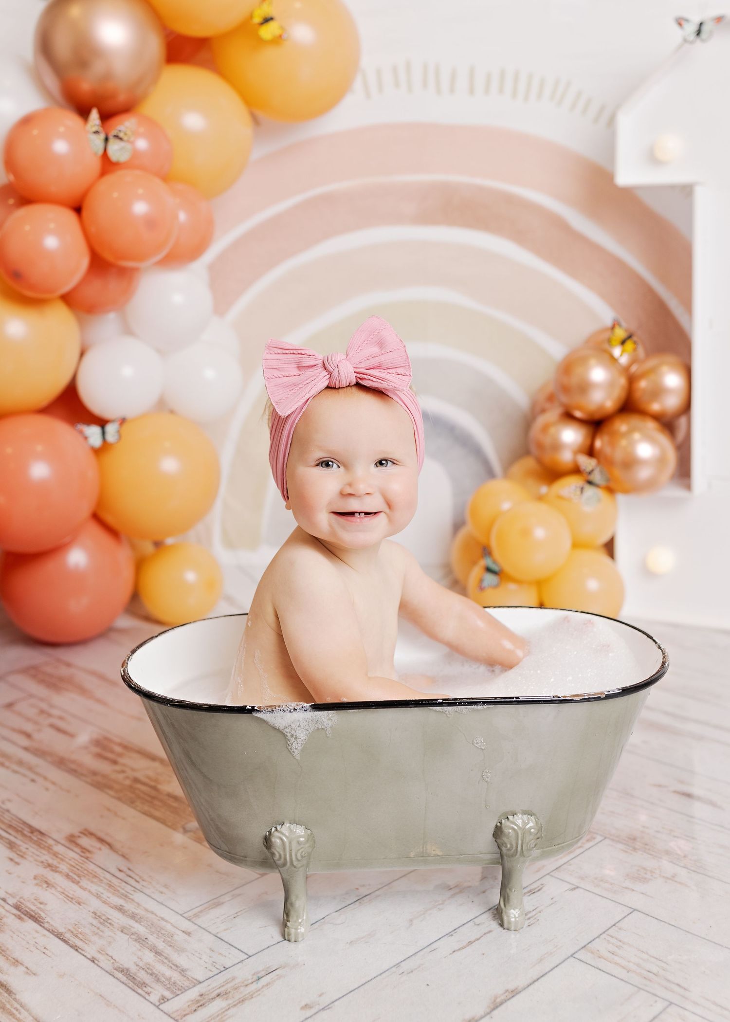 A vintage-style bathtub photo session setup with peach and gold balloons against a rustic whitewashed background.