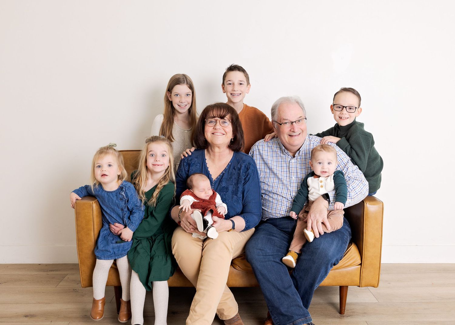 Grandparents sitting on a tan leather sofa surrounded by seven grandchildren in a bright studio family portrait.