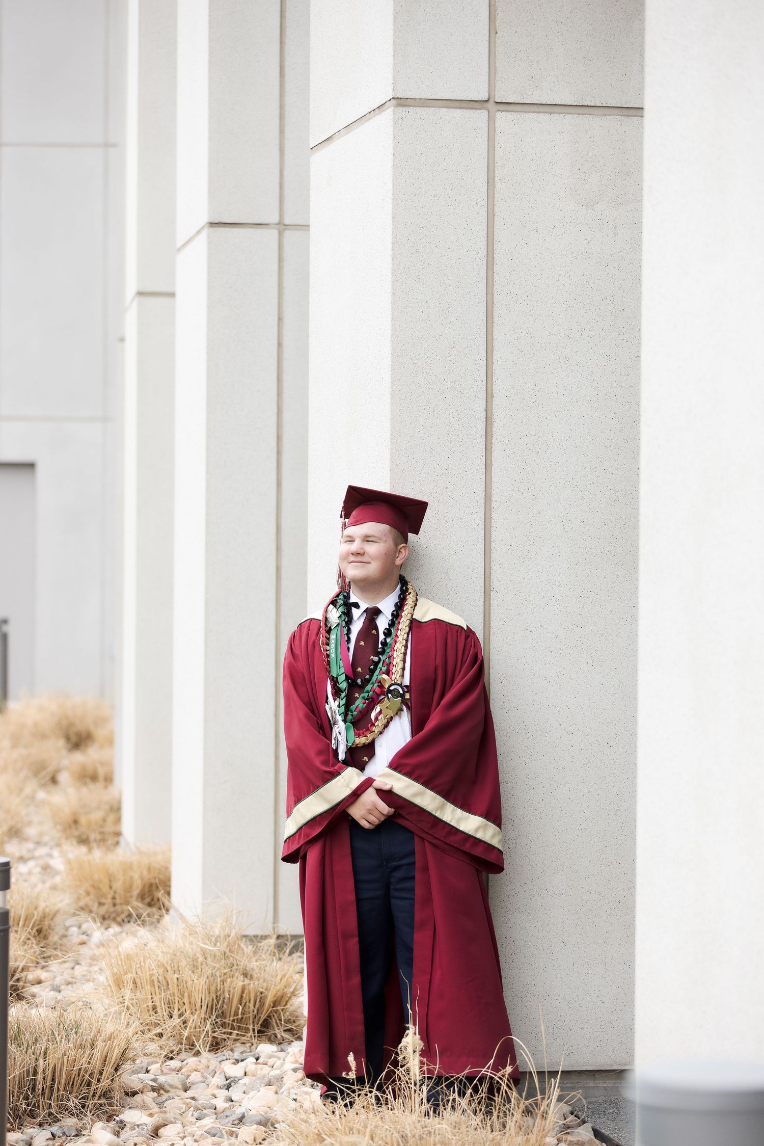 Young male graduate in maroon cap and gown with leis, leaning against white building columns outdoors.