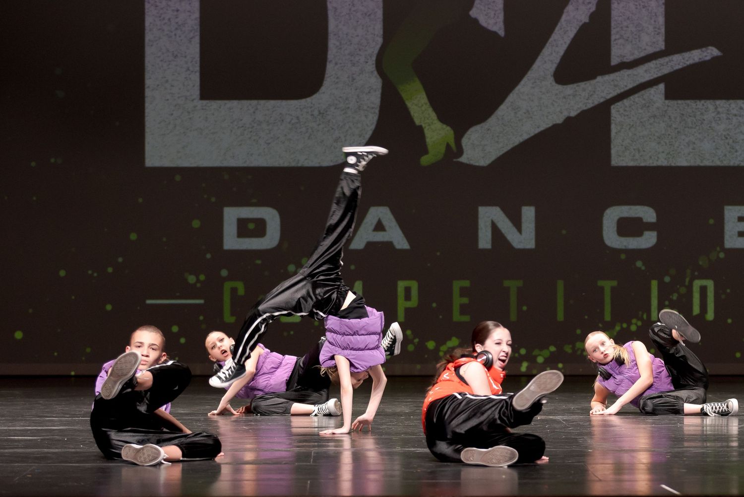 Modern dancers in black perform gracefully on a dark stage with a purple illuminated backdrop during a dance competition.