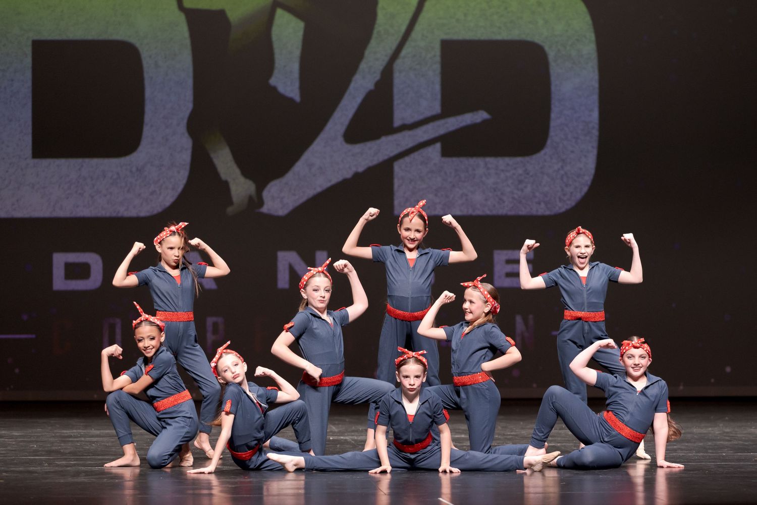 Dance performers in matching navy and red outfits strike synchronized poses on stage during a group routine.