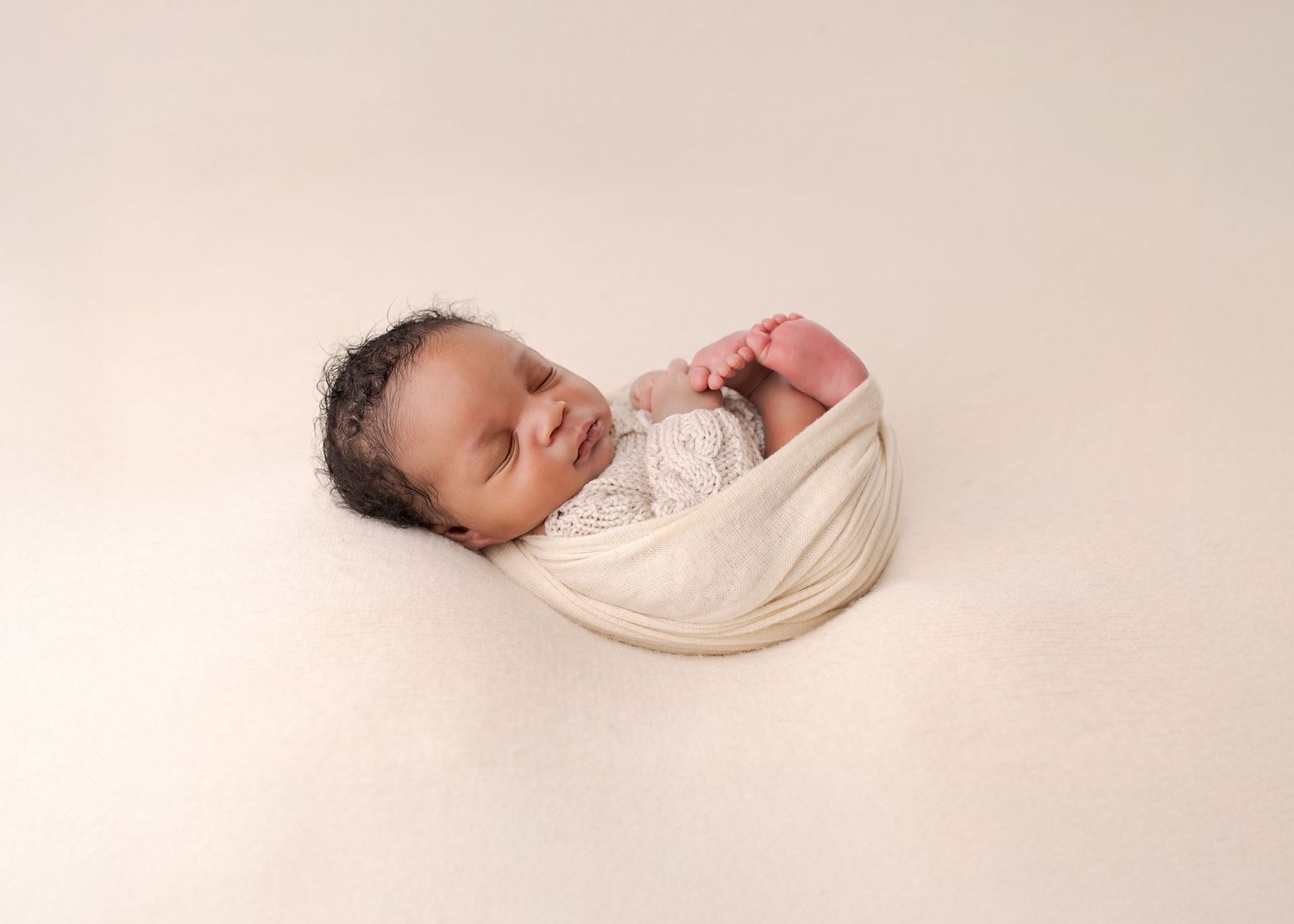 Peaceful sleeping newborn baby in soft cream tones lying on textured blanket during professional photo session.
