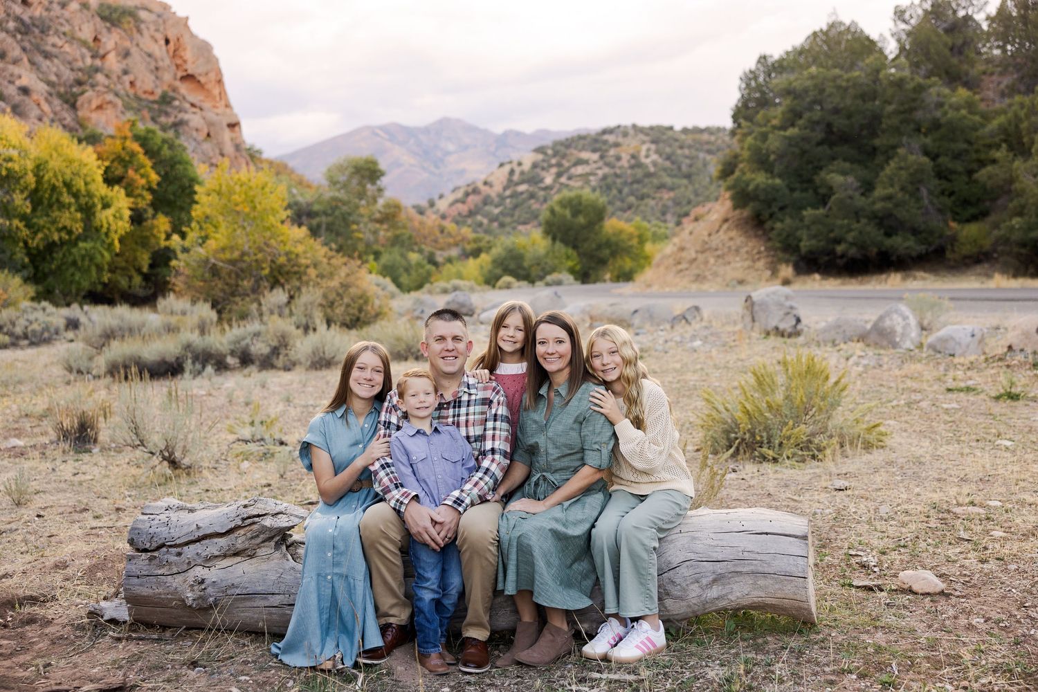 Family of six posing on a log in a scenic mountain meadow with autumn foliage and rocky peaks in the background.