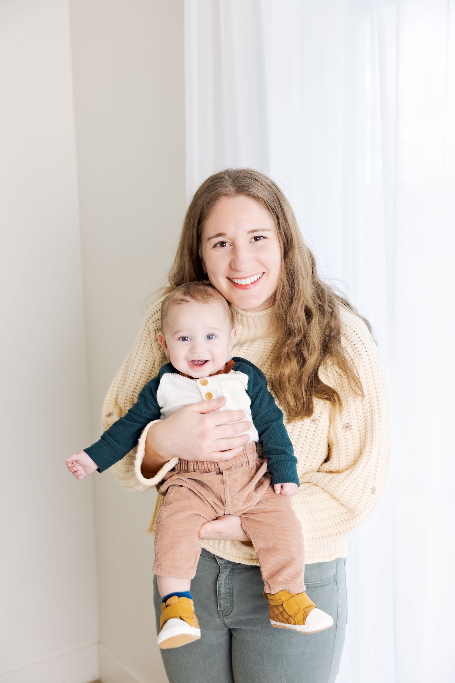 Smiling young woman in yellow sweater holding a happy baby dressed in navy and brown outfit with yellow shoes.