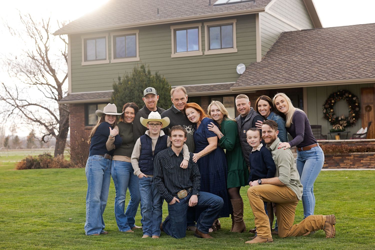 Large family group posing together outdoors in front of a two-story farmhouse on a green lawn.