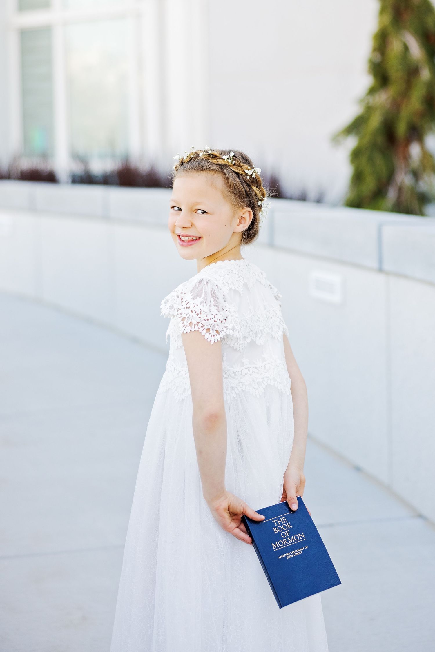 A person in a white lace dress holds a blue book while standing against a white building.