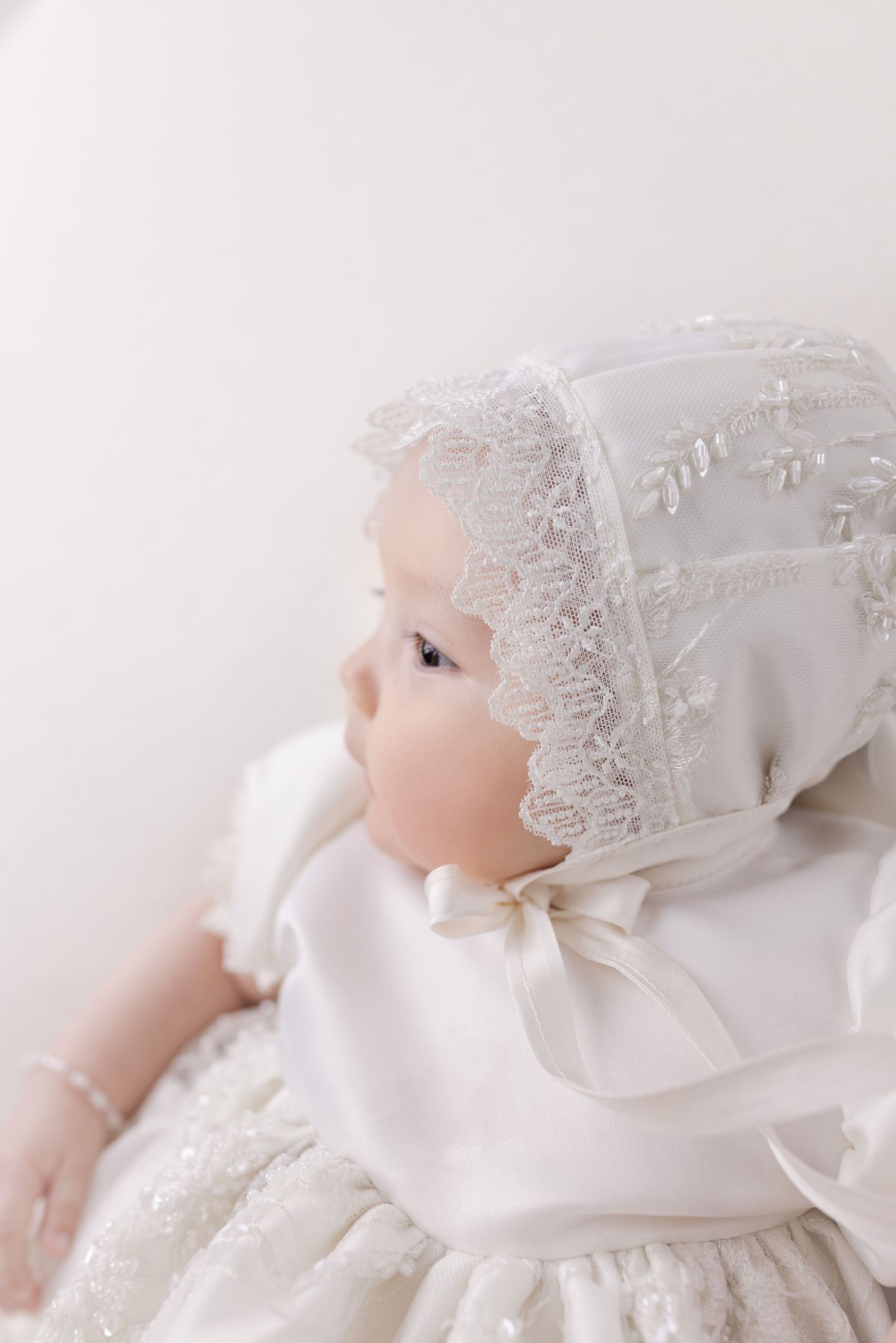 Baby in white christening gown with delicate lace bonnet in profile view.