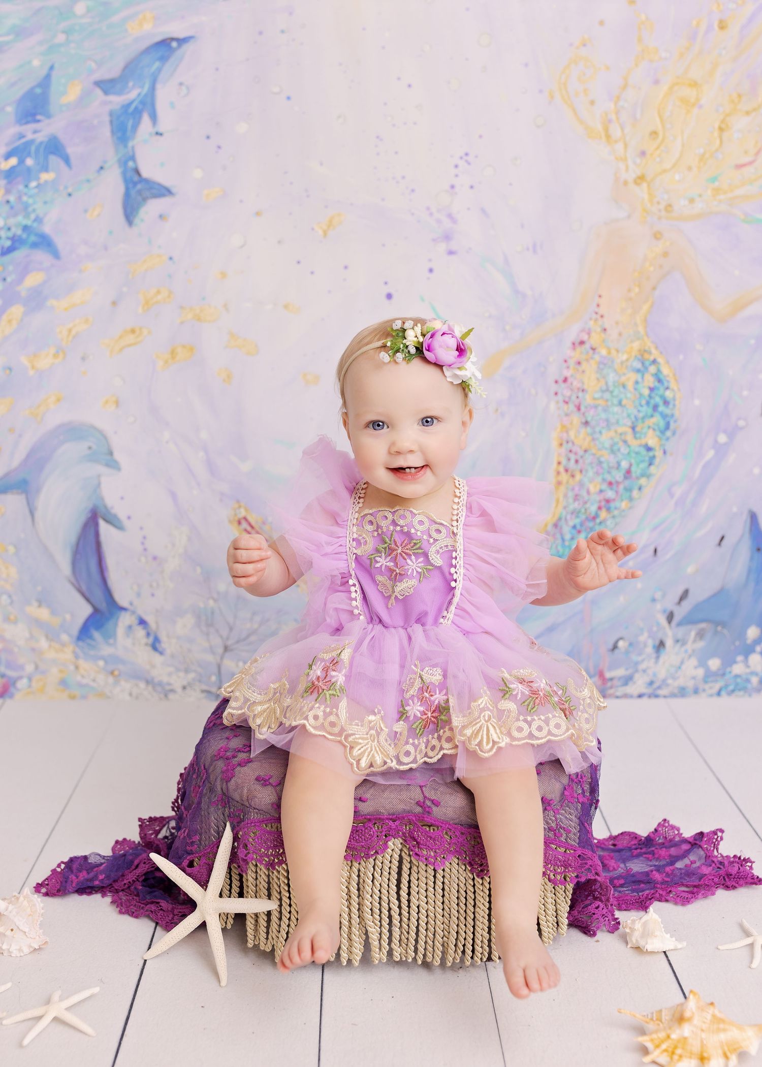 Sweet young child in pink tulle dress sits on purple decor with starfish and ocean-themed backdrop.