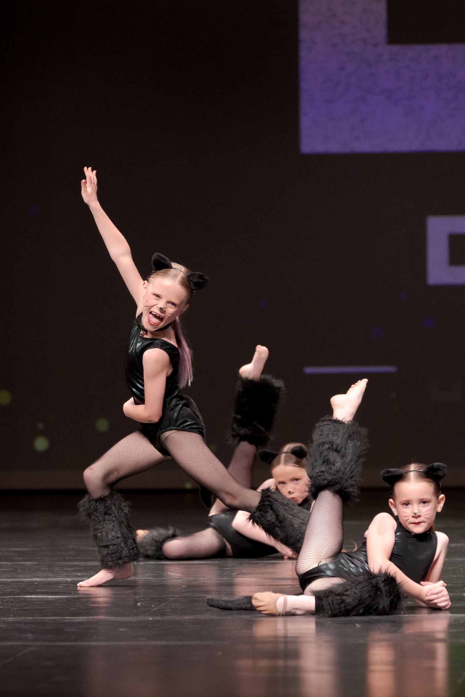 Young dancers in black outfits performing dynamic choreography on a dark stage with graceful poses and movements.