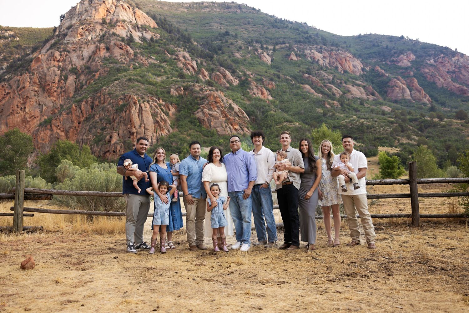 Large family group portrait outdoors with red rock mountains and green trees in background on a sunny day.
