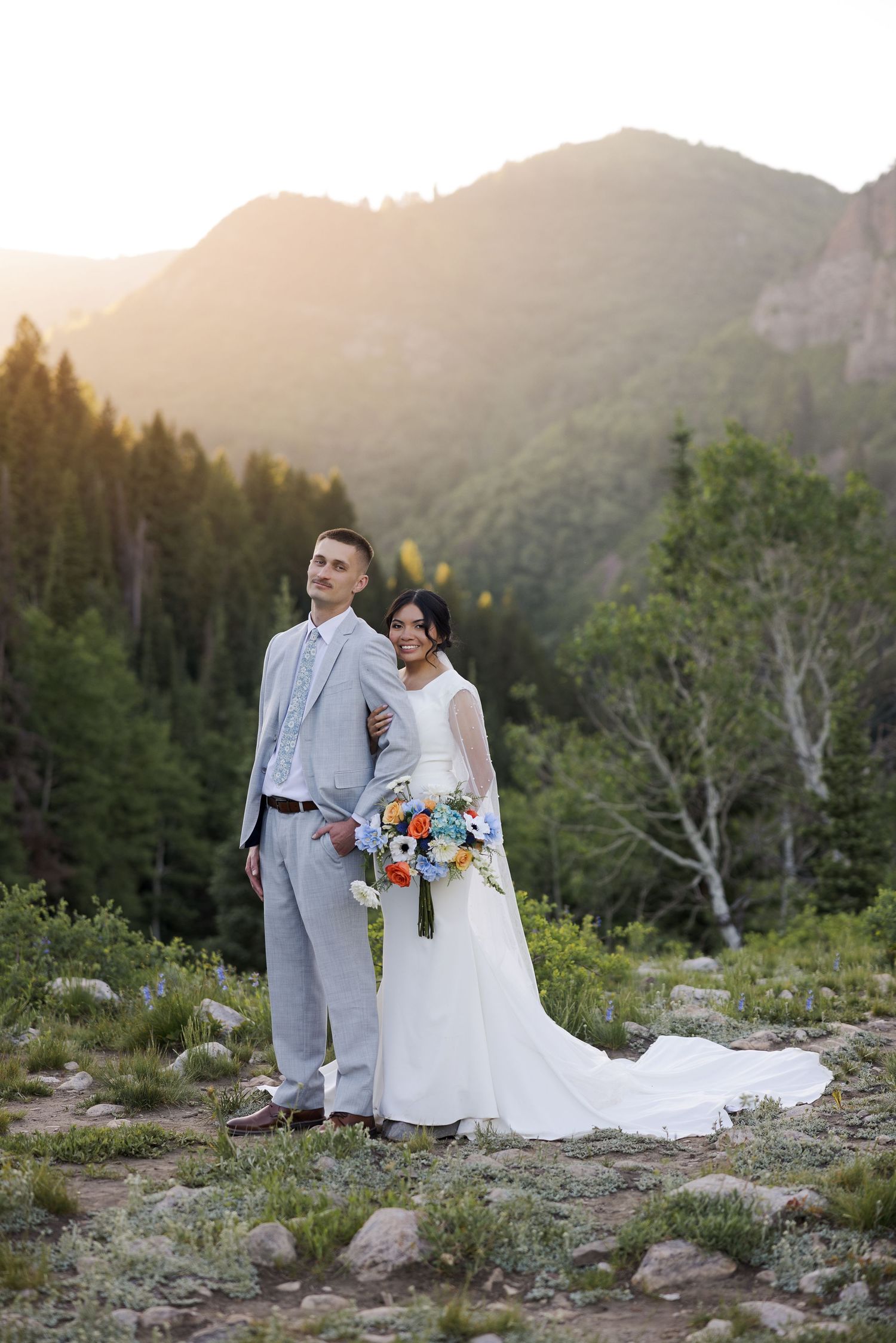 Wedding couple poses against a stunning mountain backdrop at sunset wearing white and light gray attire.