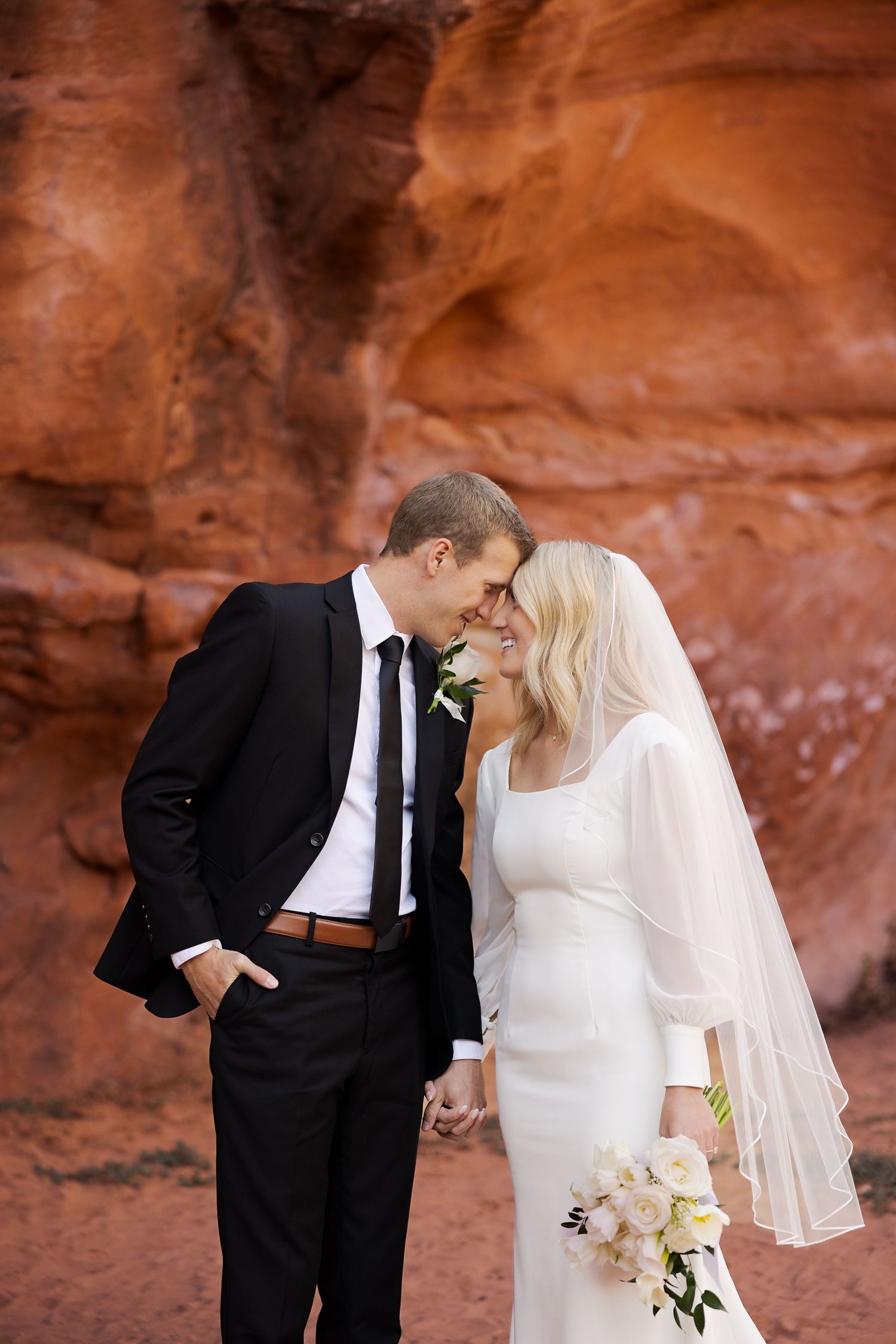 Elegant wedding couple shares intimate moment against red sandstone cliffs.