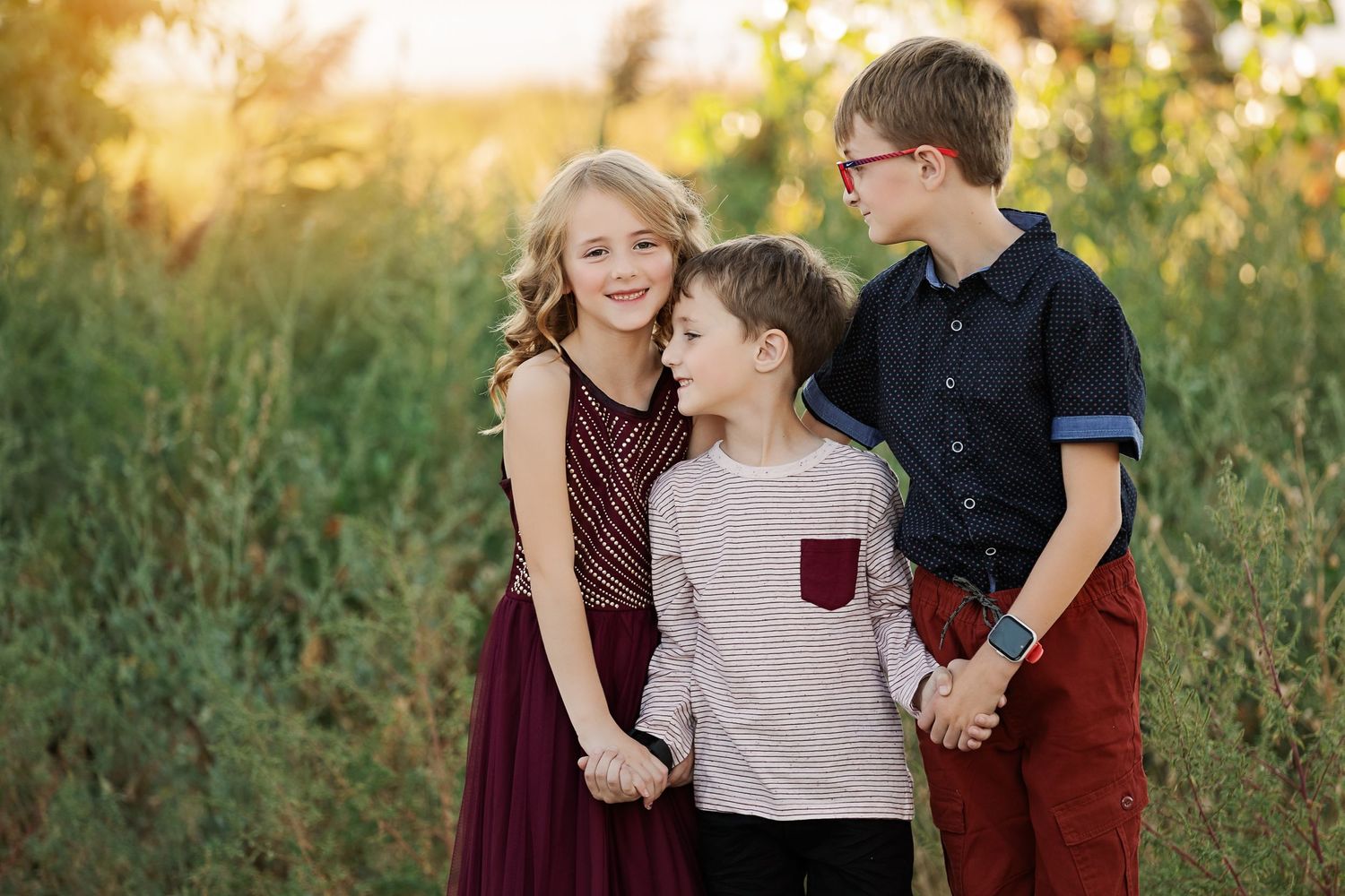 Three children stand together outdoors at sunset in coordinated casual outfits.
