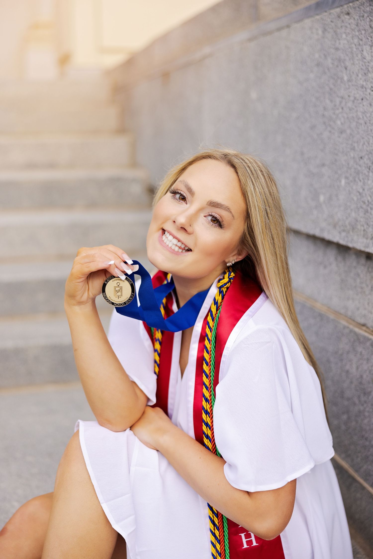 Graduate in white shirt and graduation regalia holds up gold medal during outdoor portrait session.