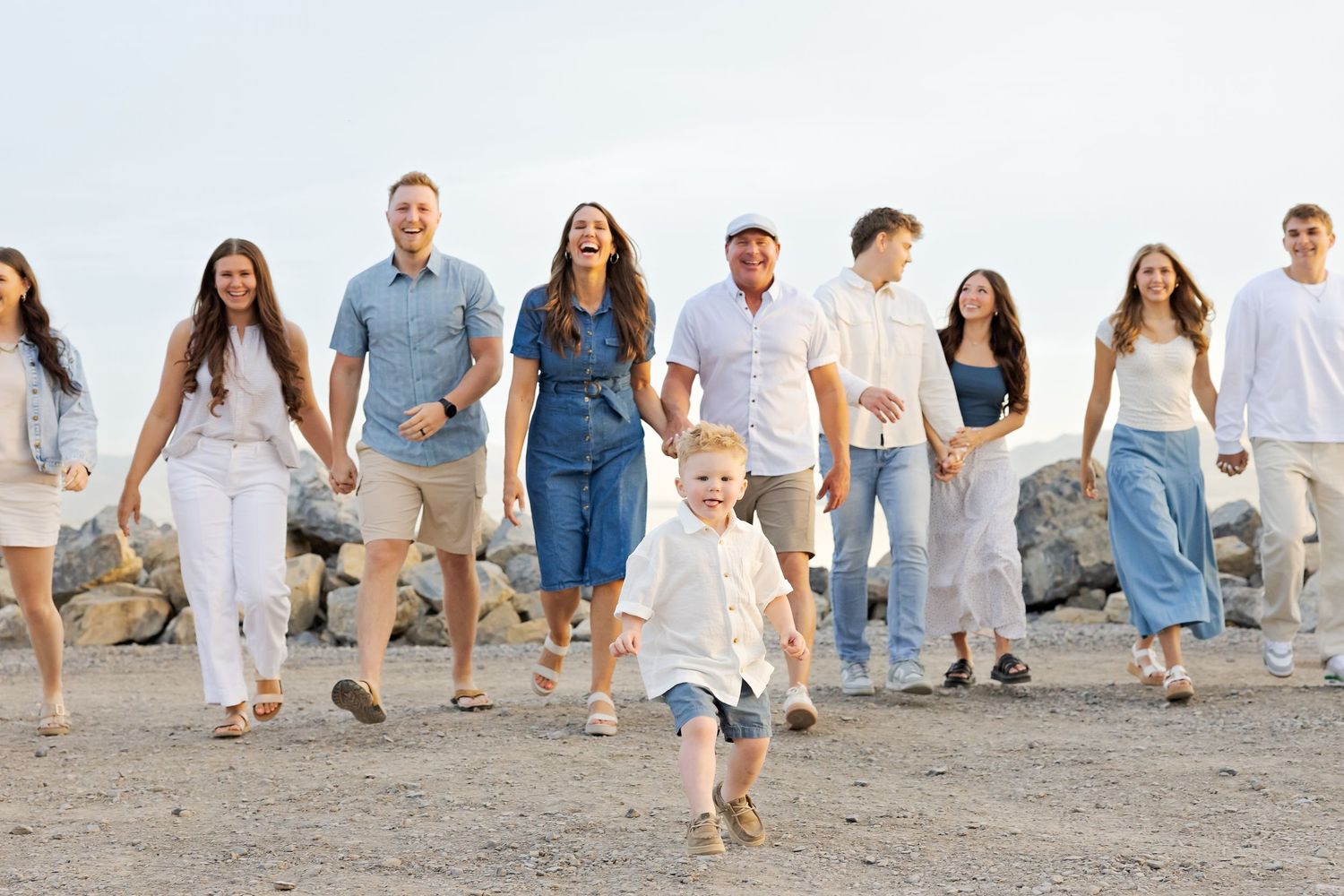 Large family walking together on a rocky beach, smiling and holding hands, with a young child running ahead.