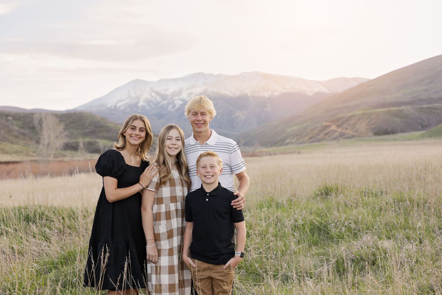 Family of four smiling in a golden field with mountain backdrop at sunset.