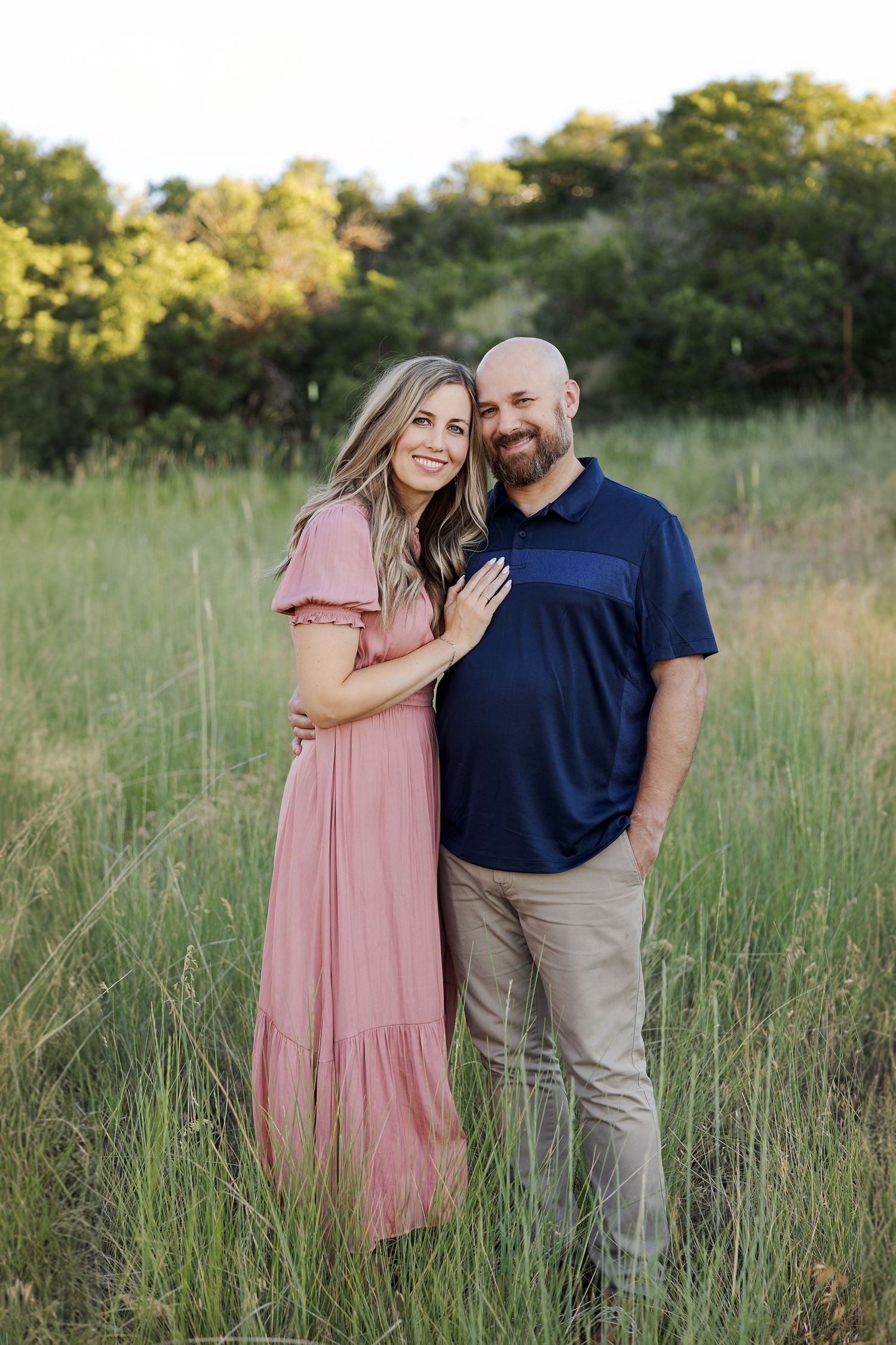 Couple in casual attire embracing in tall grass during sunset.