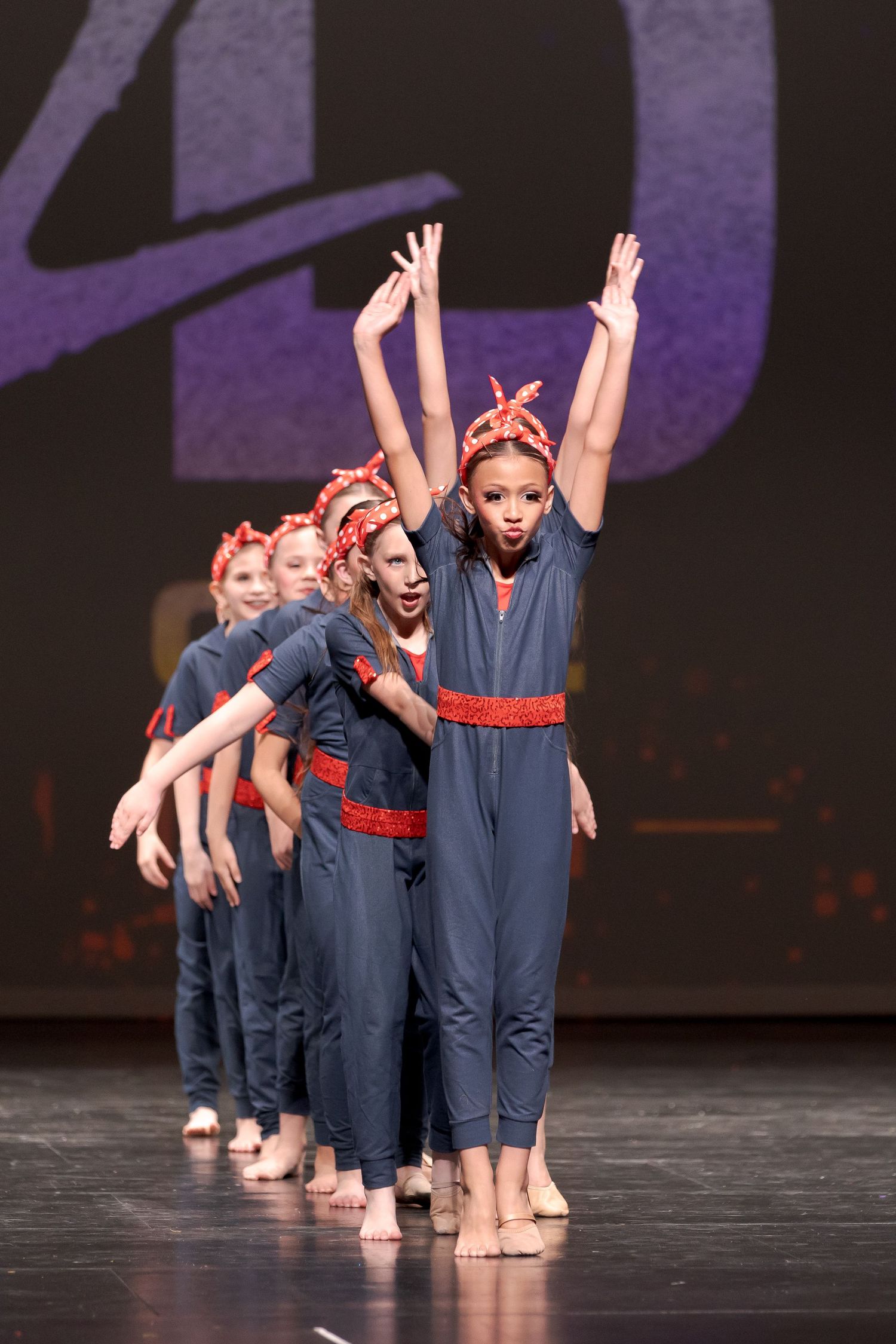 Dancers in navy blue outfits perform a synchronized dance routine on a dark stage with dramatic lighting.
