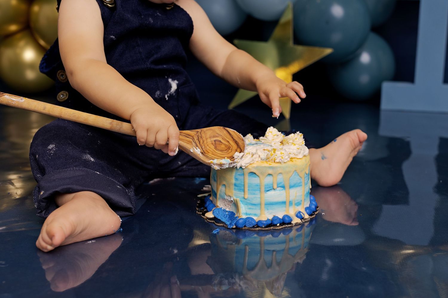 A toddler's hands reach for a blue and white decorated birthday cake with candles on a dark table surface.