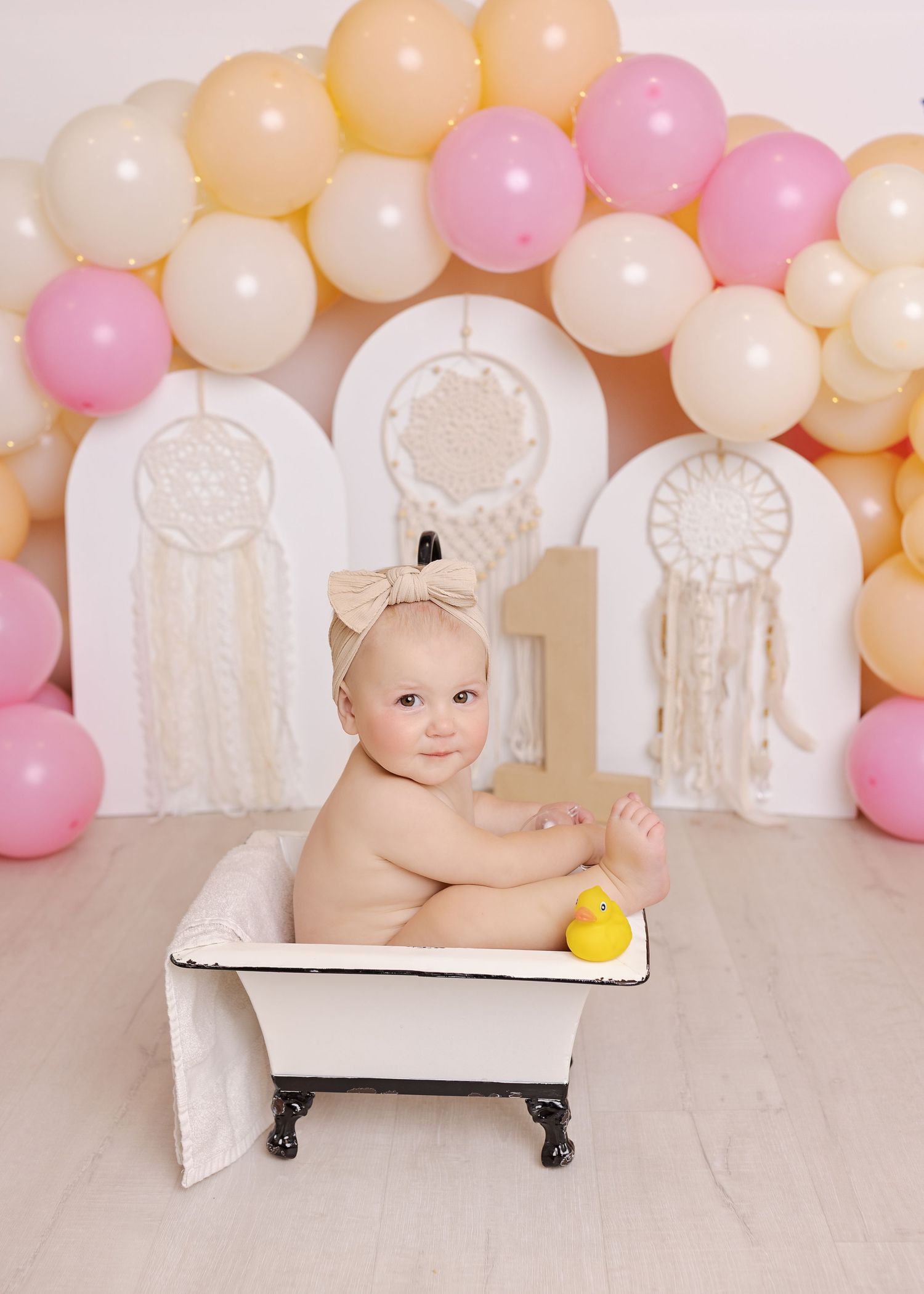A baby sits in a white vintage bathtub surrounded by pink and peach balloons for a whimsical photo session.