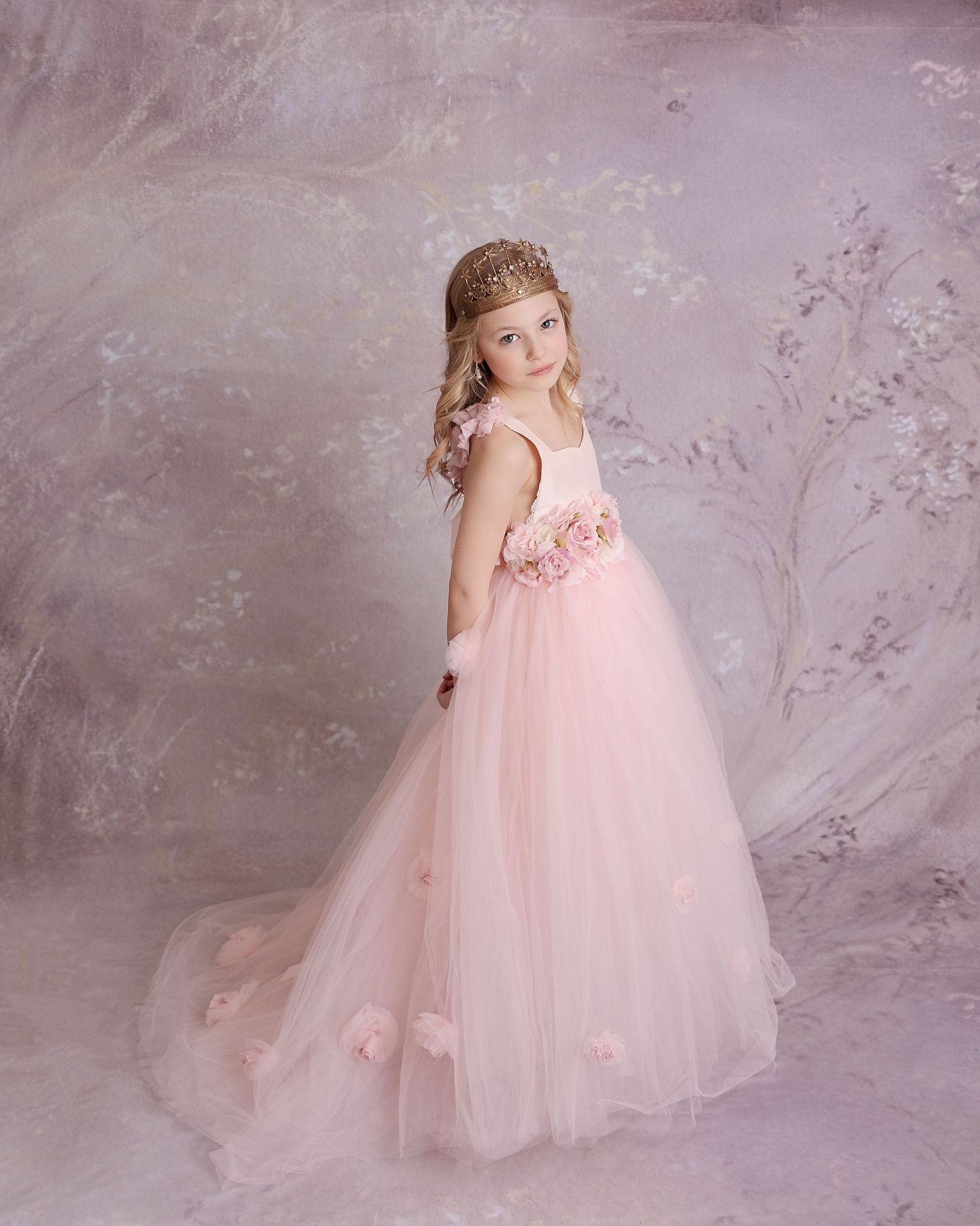 A child wearing a flowing pink princess dress and tiara poses against a soft gray background.