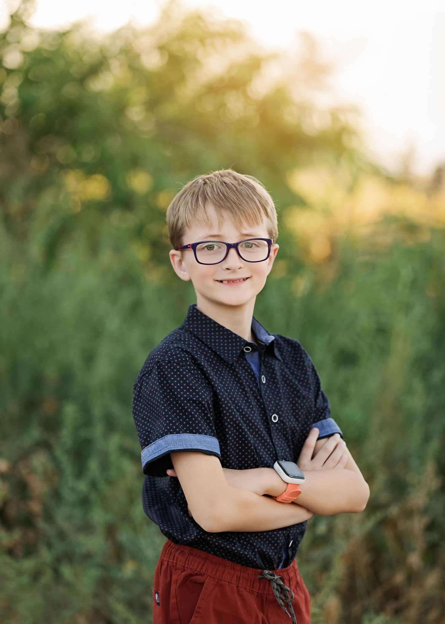 Young person in polka dot shirt and glasses poses confidently against green summer background.