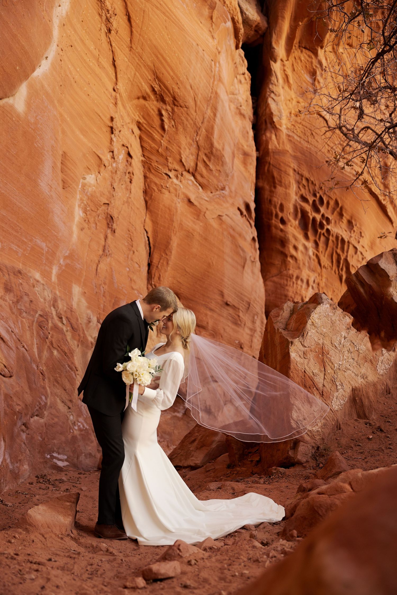 Wedding portraits in dramatic red rock canyon with flowing white veil and classic black suit.