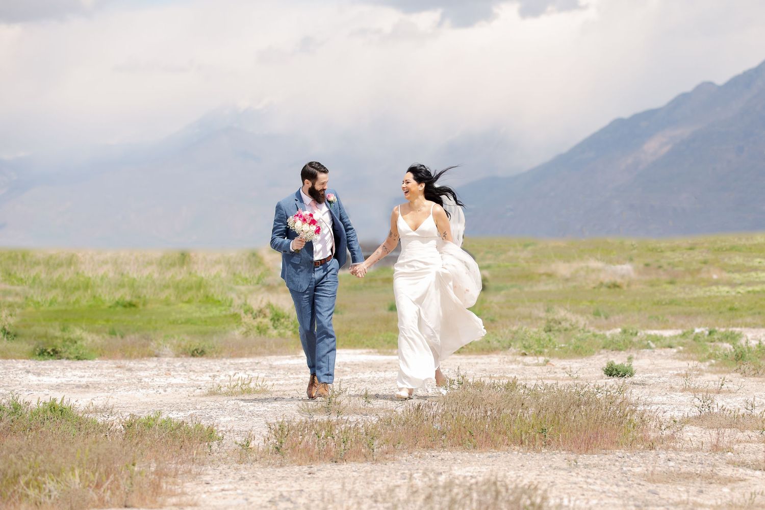 Joyful wedding couple running through a mountain meadow with dramatic cloudy sky.