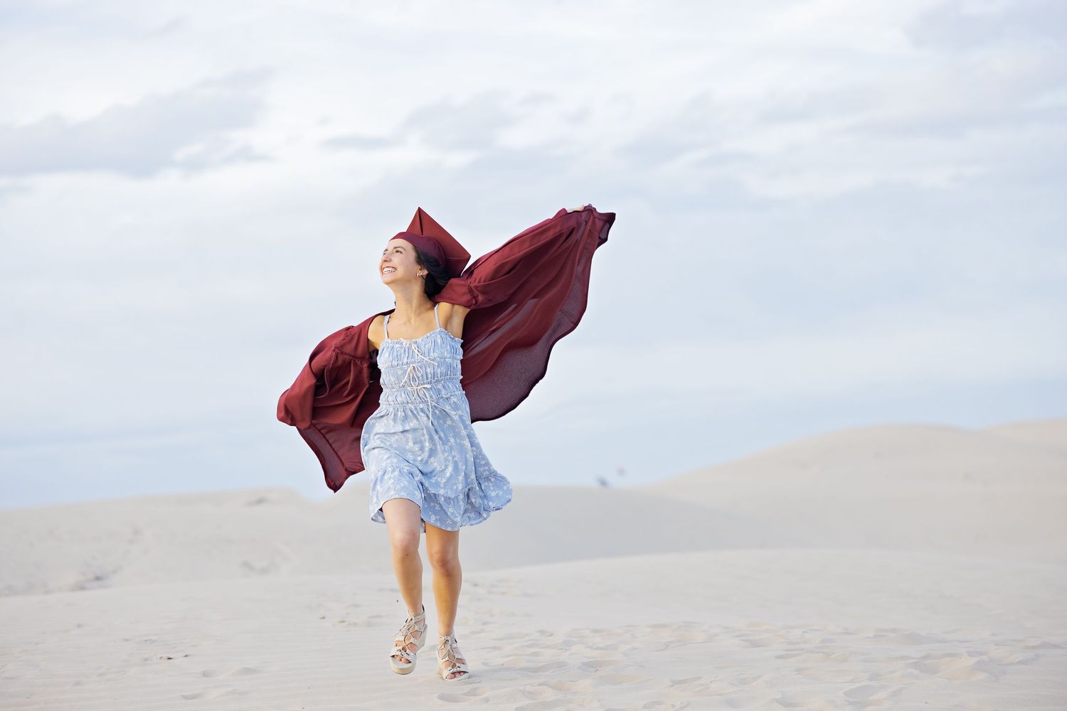A joyful figure in a blue dress twirls on a sandy beach with a flowing burgundy shawl caught in the wind.