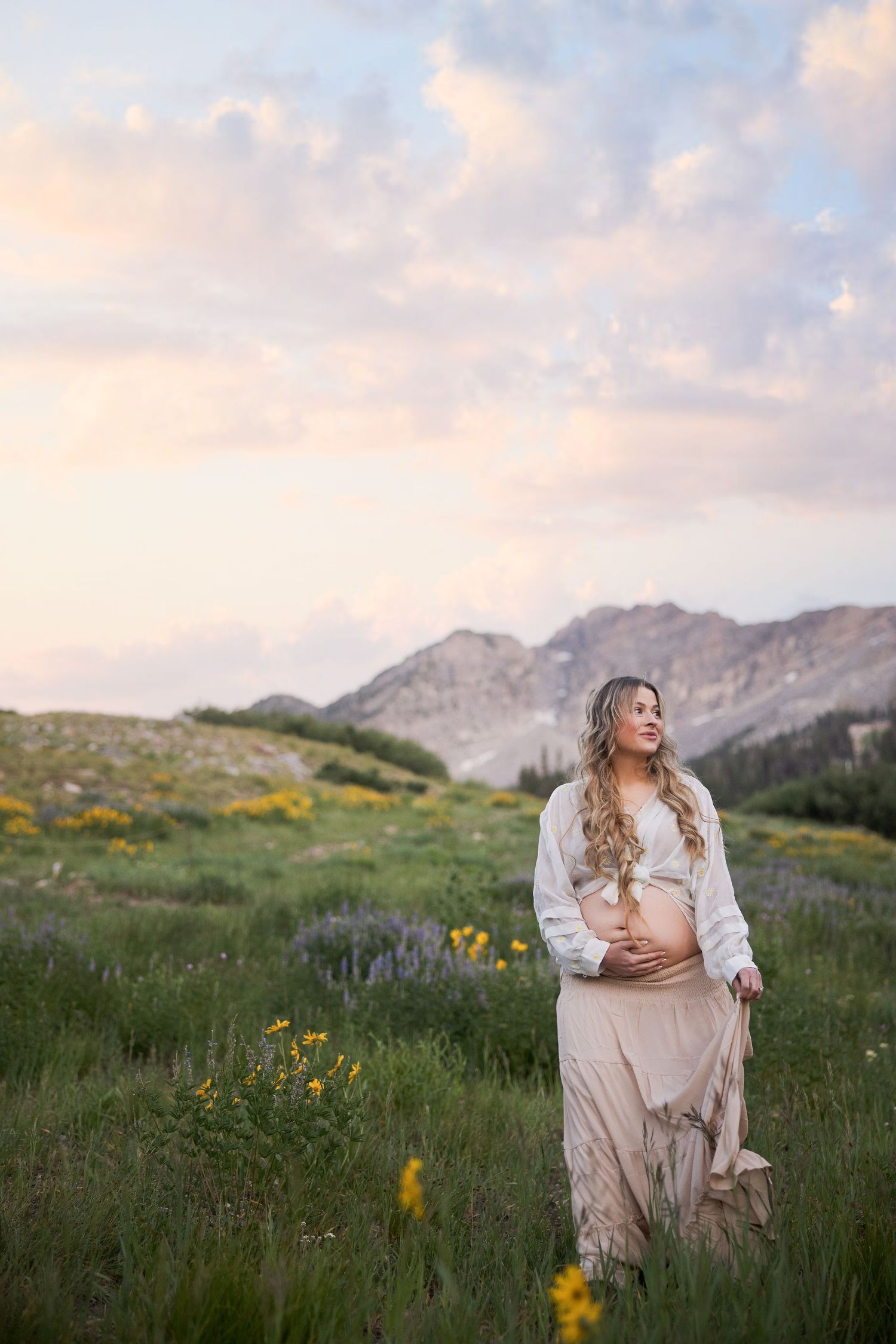 A pregnant woman in flowing dress stands in a mountain meadow with wildflowers at sunset in the wilderness.