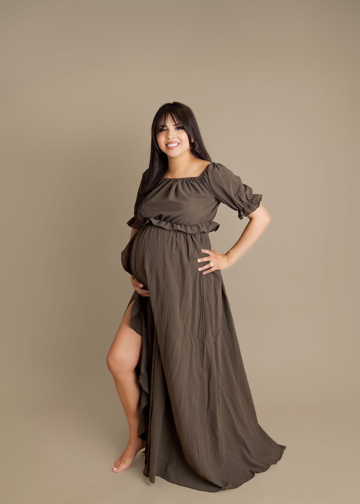 Pregnant woman in olive green off-shoulder maternity gown with flowing skirt poses gracefully in studio.
