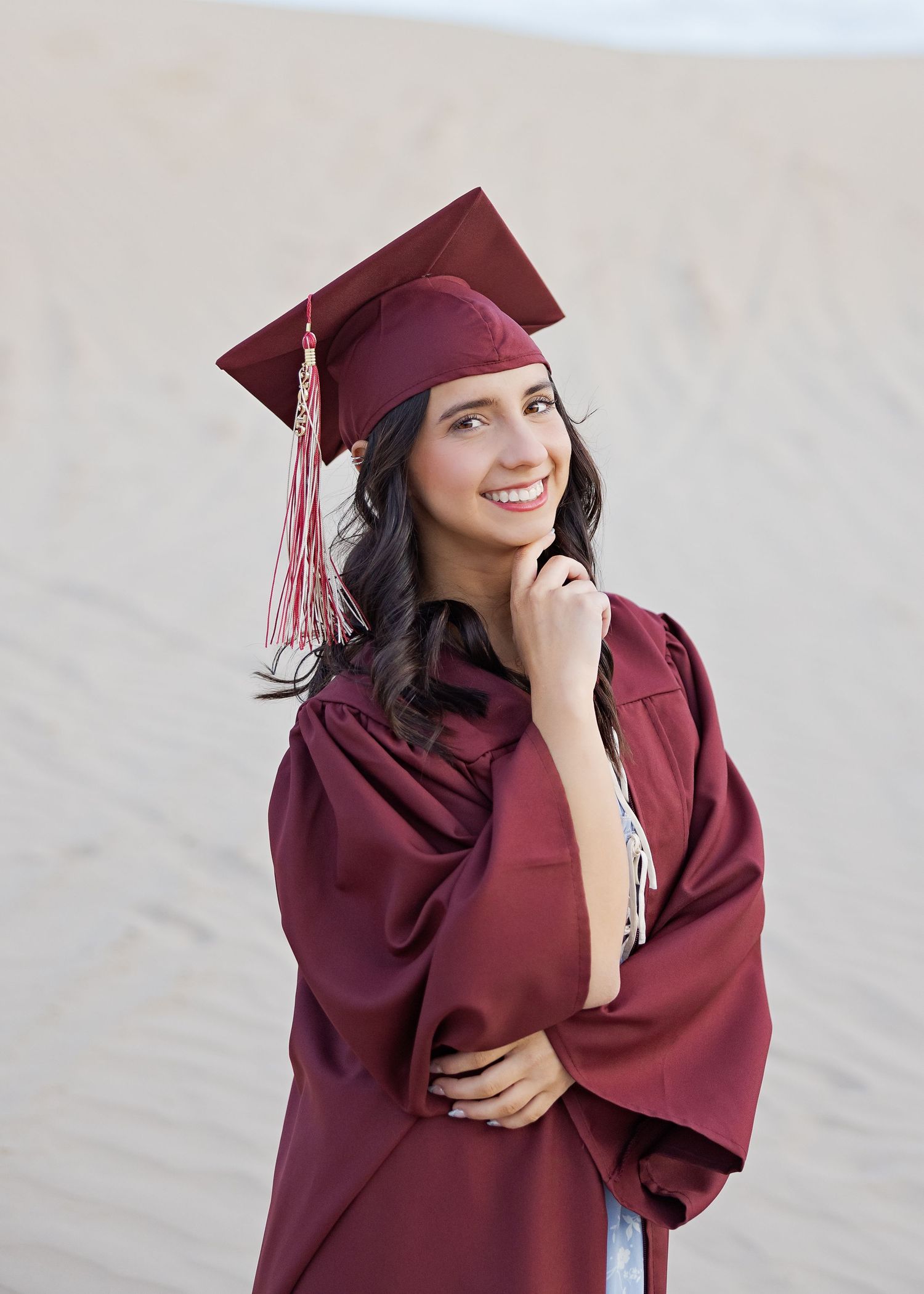 Graduate in burgundy cap and gown poses thoughtfully against neutral background.