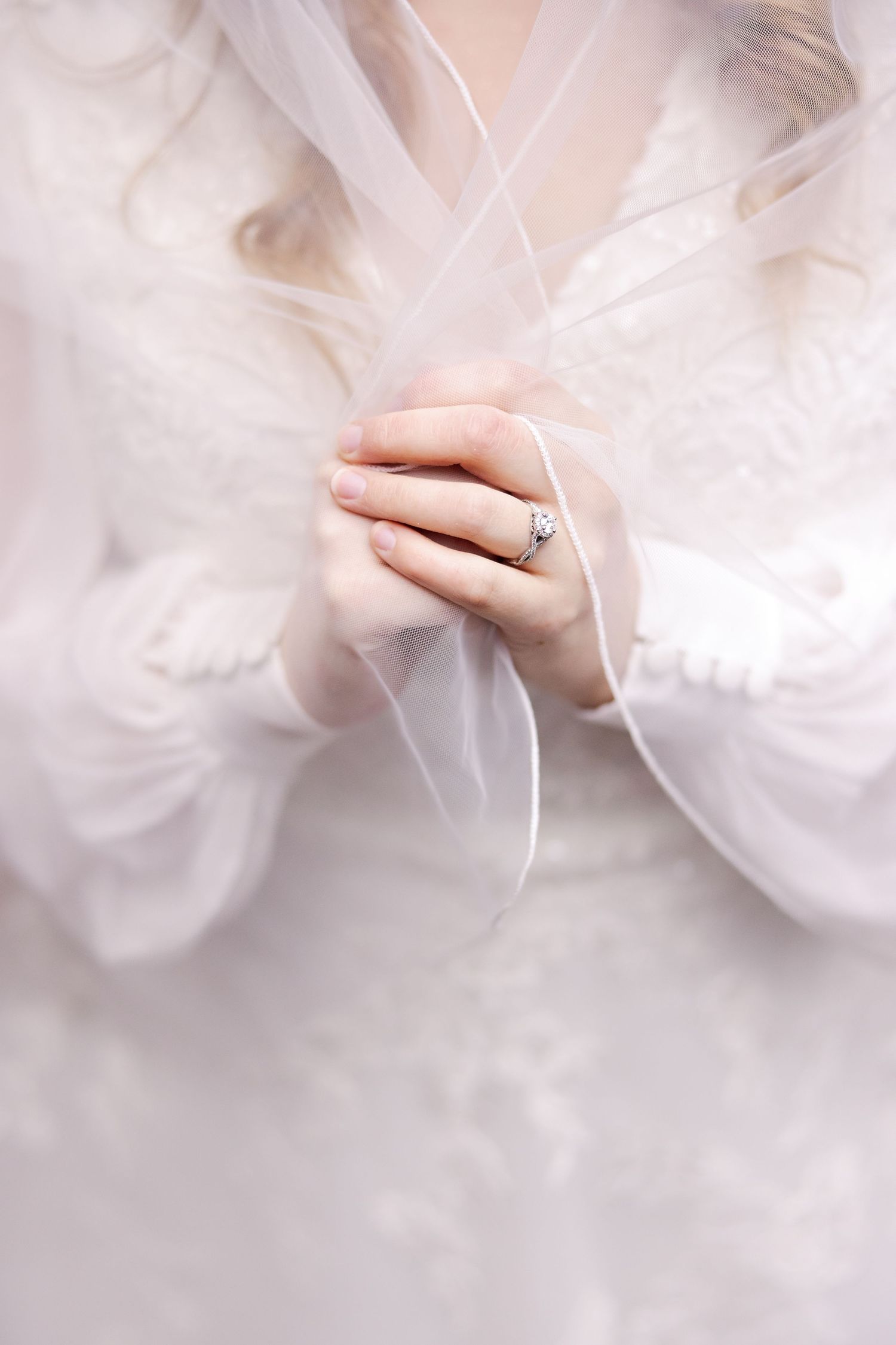 Delicate close-up of hands wearing engagement ring through sheer white veil fabric.