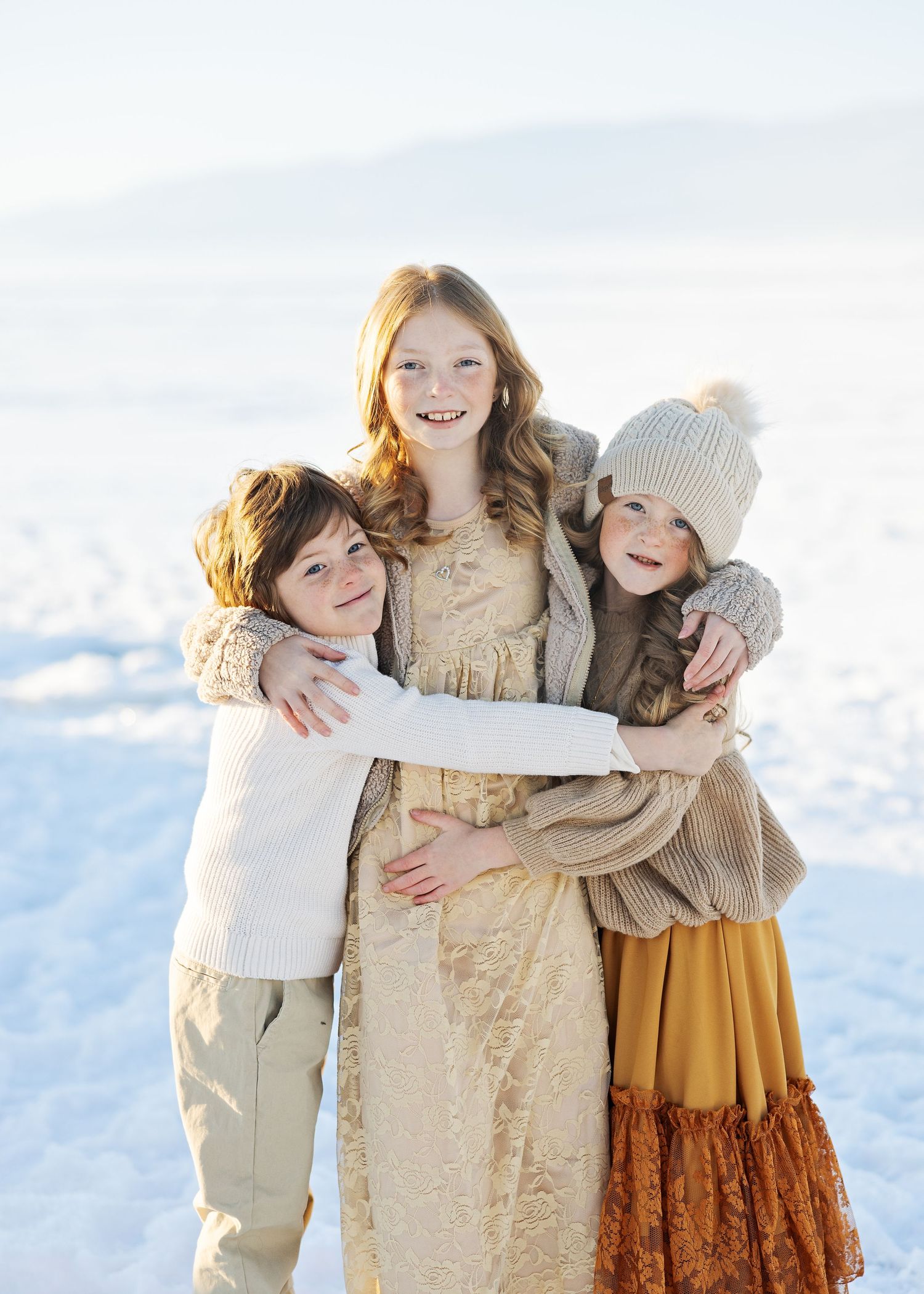 Three siblings hug and smile together in a snowy winter landscape wearing warm knit clothing.