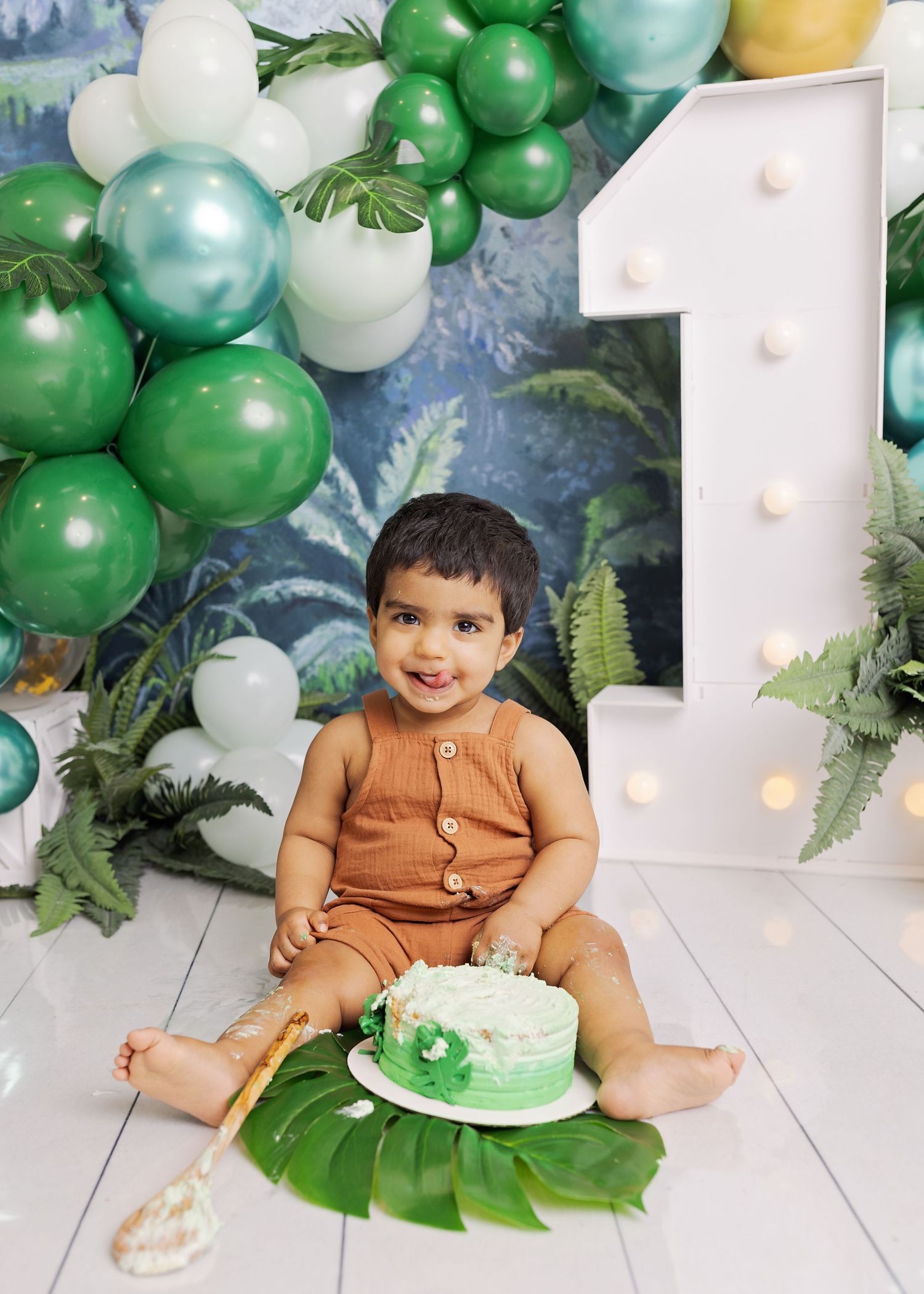 First birthday celebration setup with green and white balloons, glowing number 1 sign, tropical leaves and small cake.