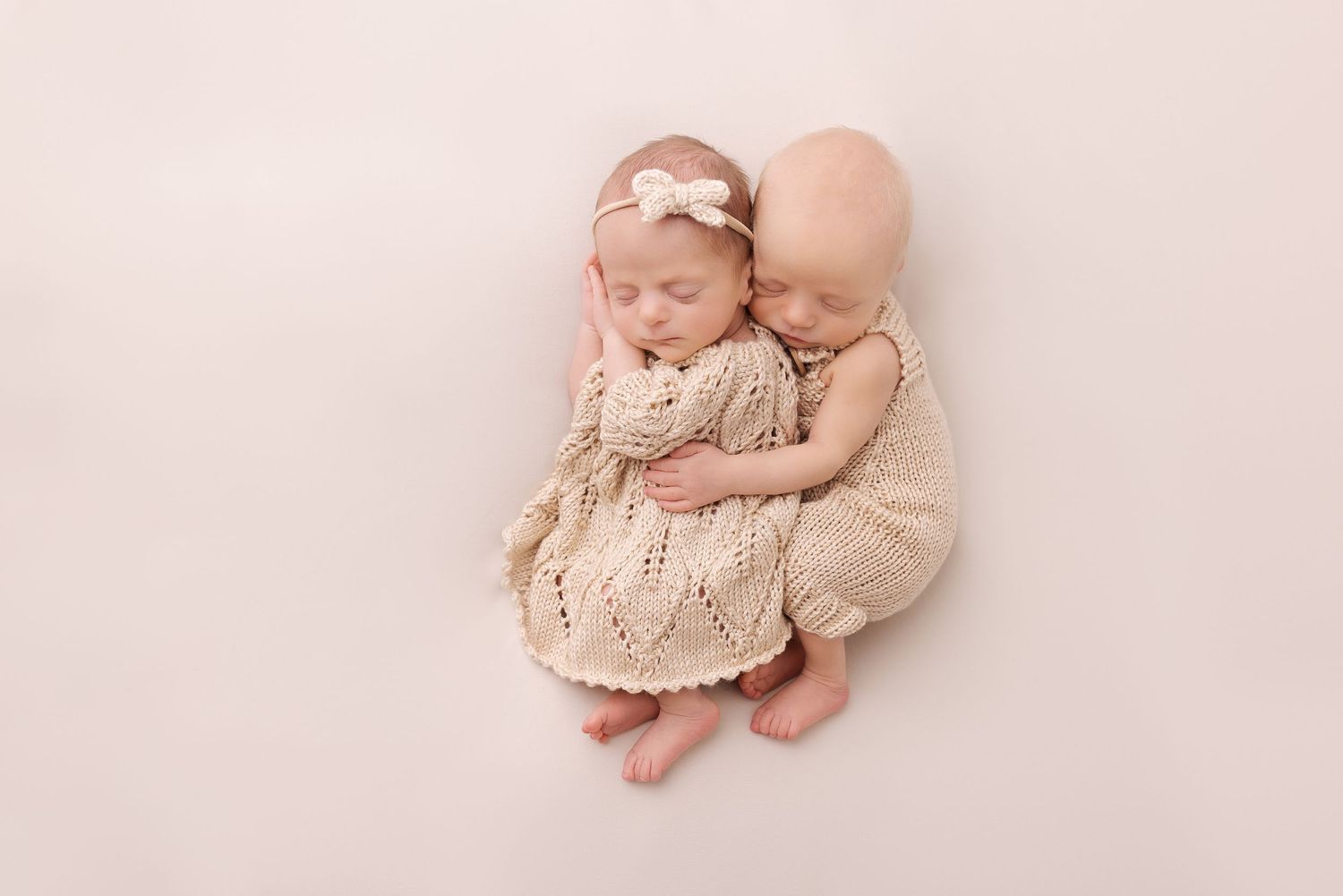 Newborn dressed in cream knit dress lying curled up on soft beige backdrop for portrait session.