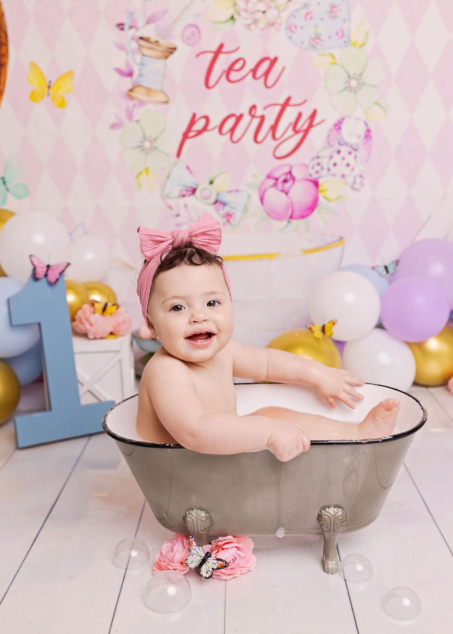 Whimsical tea party themed birthday setup with pastel balloons, butterflies and decorative metal bathtub against pink backdrop.