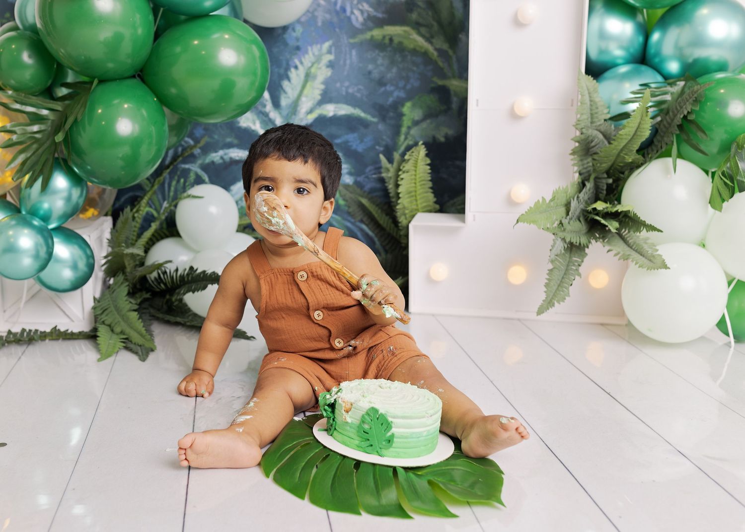 Tropical cake smash photo session setup with green and white balloons, palm leaves, and a one year old eating cake.