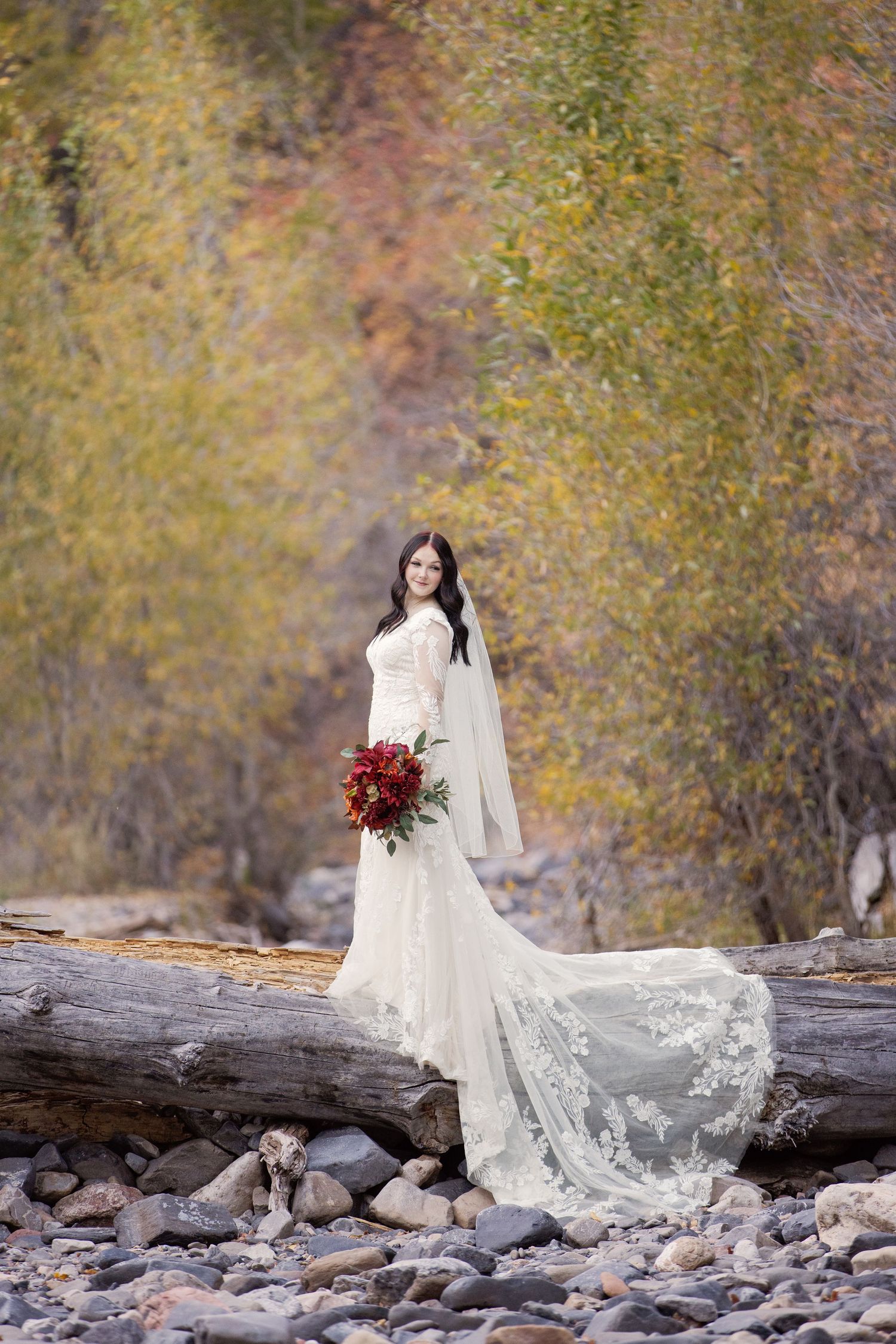 Bride with lace gown and burgundy bouquet posed on rocky riverbed with autumn foliage.