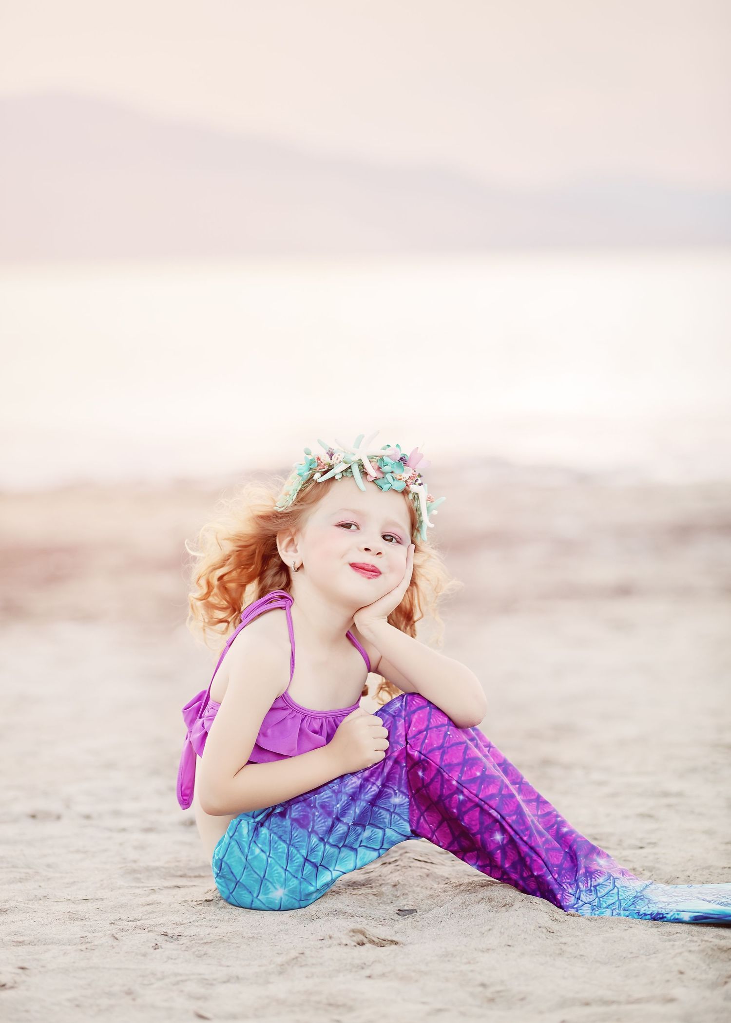 A beachside portrait of someone in a purple and blue mermaid costume with seashell crown.