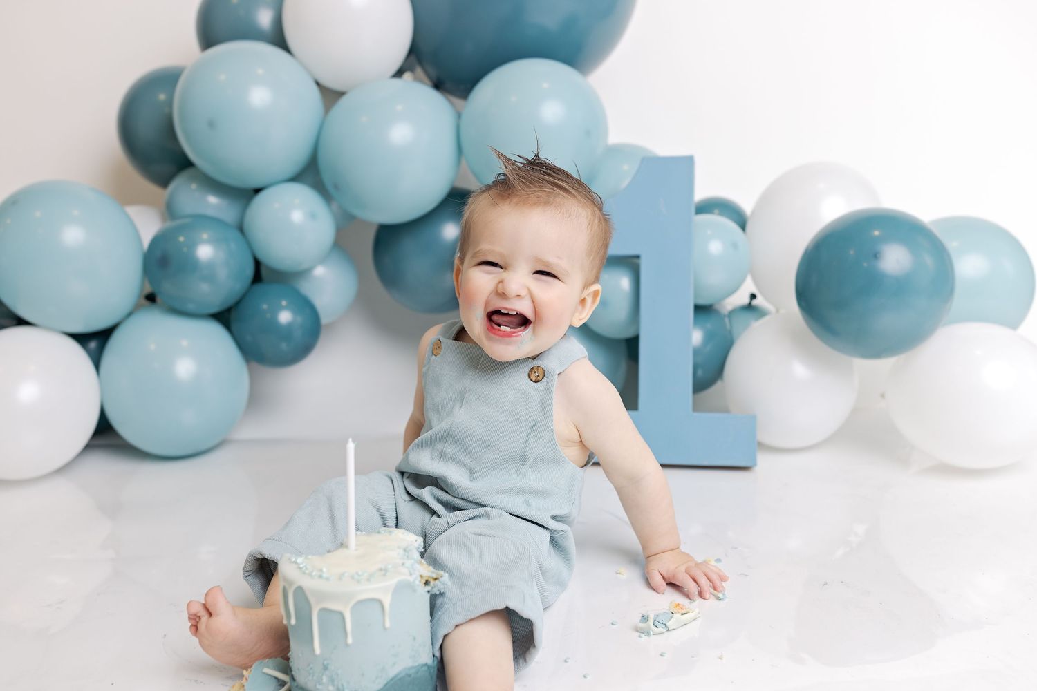 Blue and white balloon arch decorates a first birthday celebration with cake and overjoyed expression.