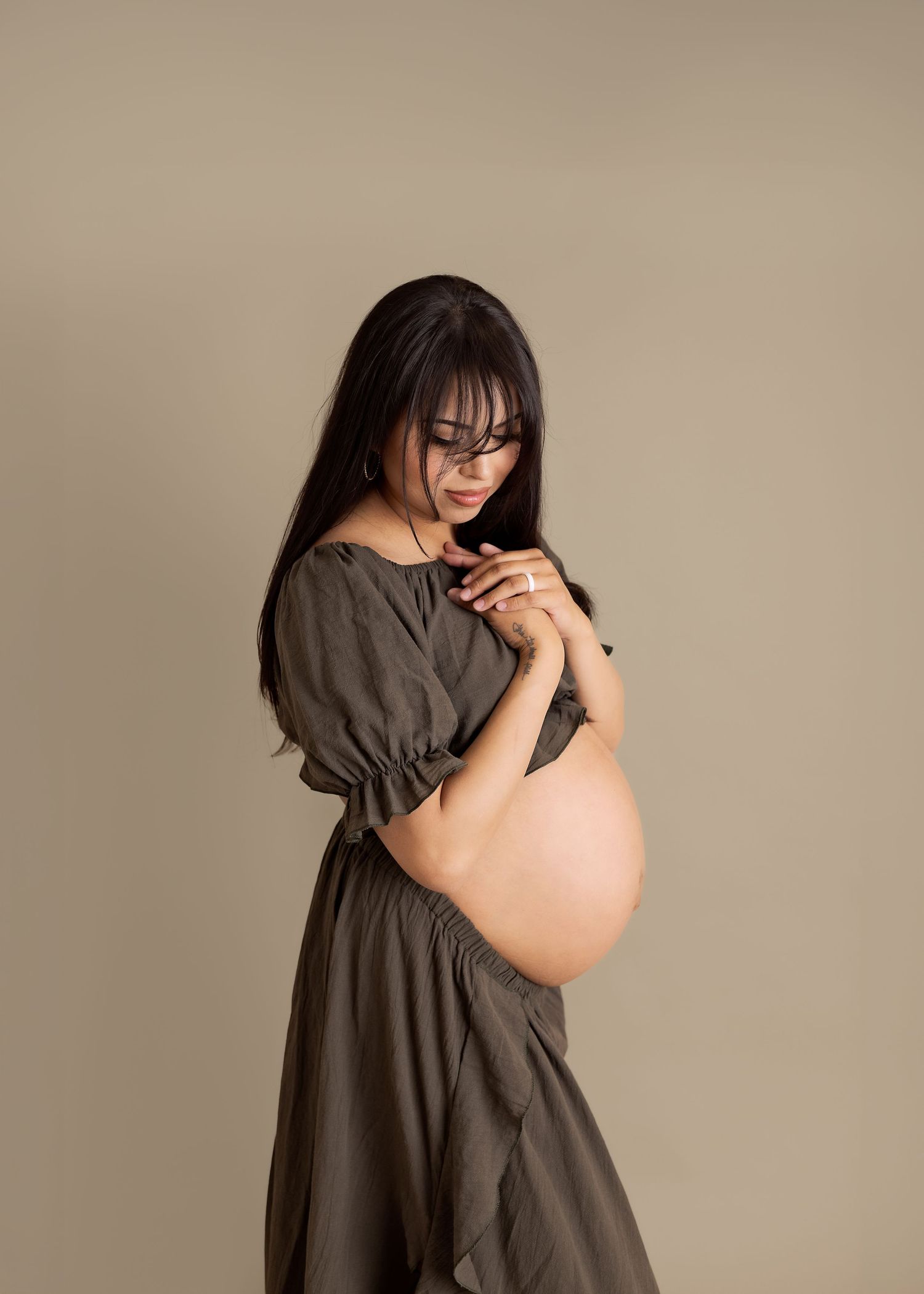 Pregnant woman in olive green dress with exposed belly, wearing glasses, posing against beige background.